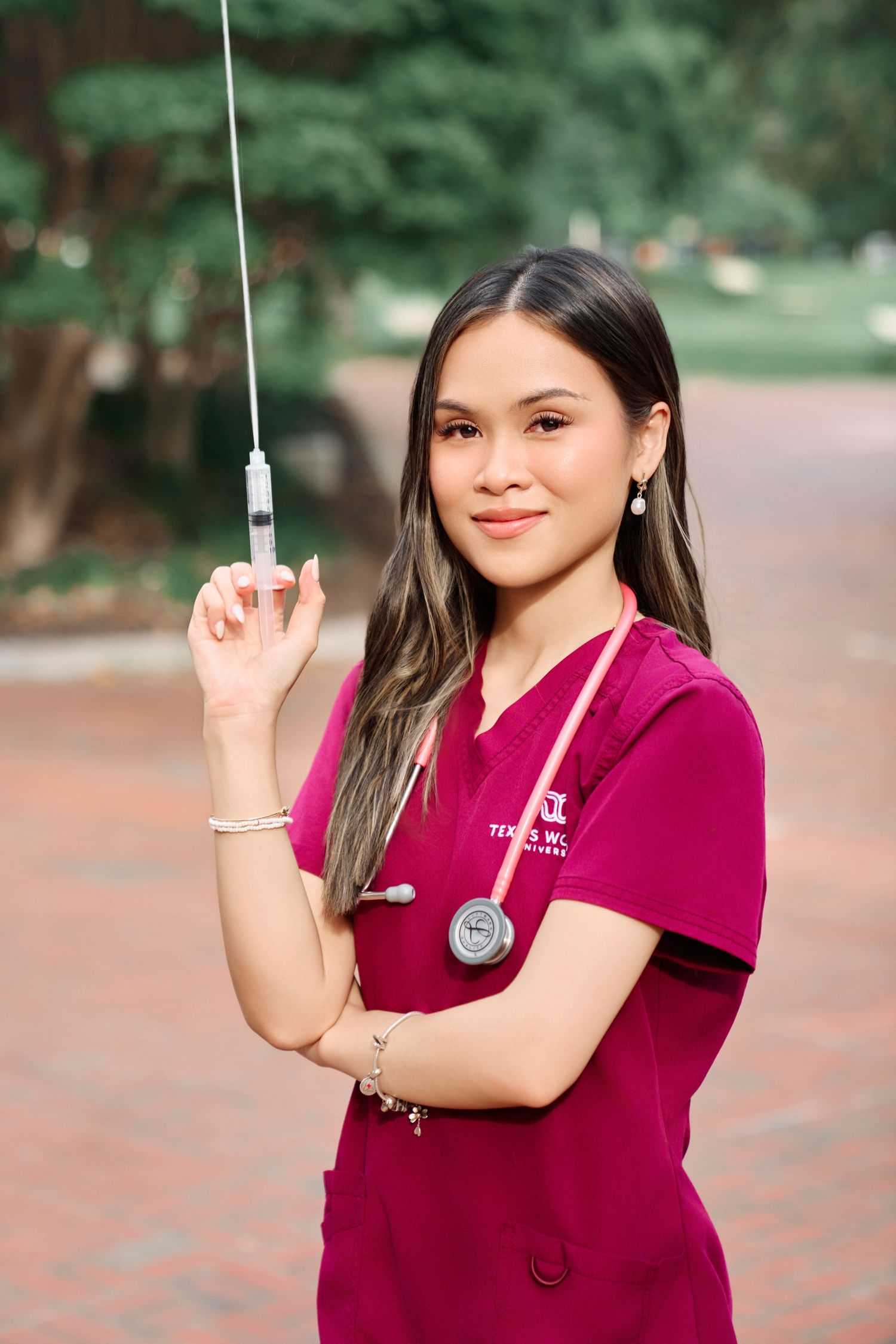 Young nurse in magenta scrubs holding a syringe upright while wearing a stethoscope outdoors.