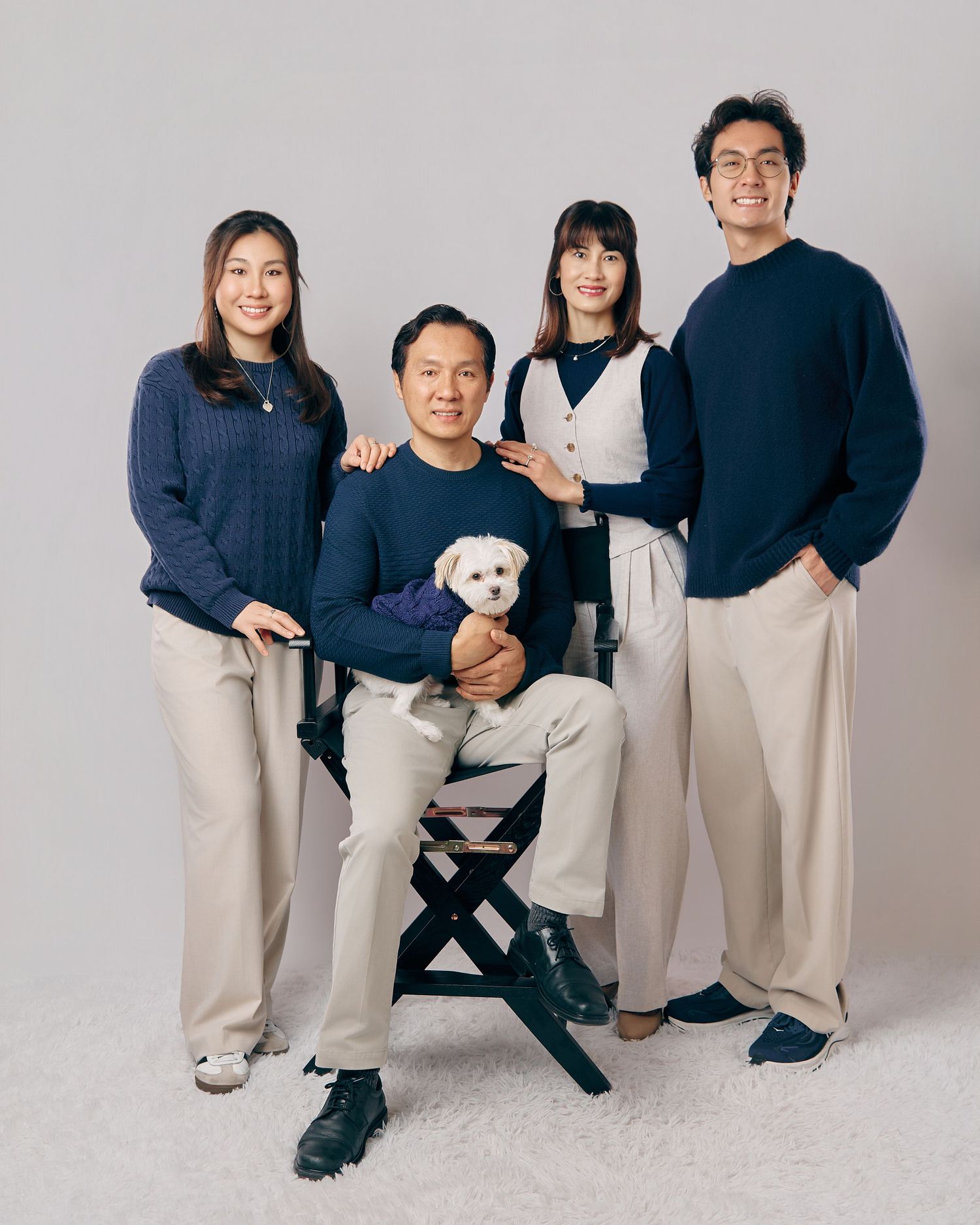 Asian family of four posing in coordinated navy and beige outfits with a white dog in a studio.