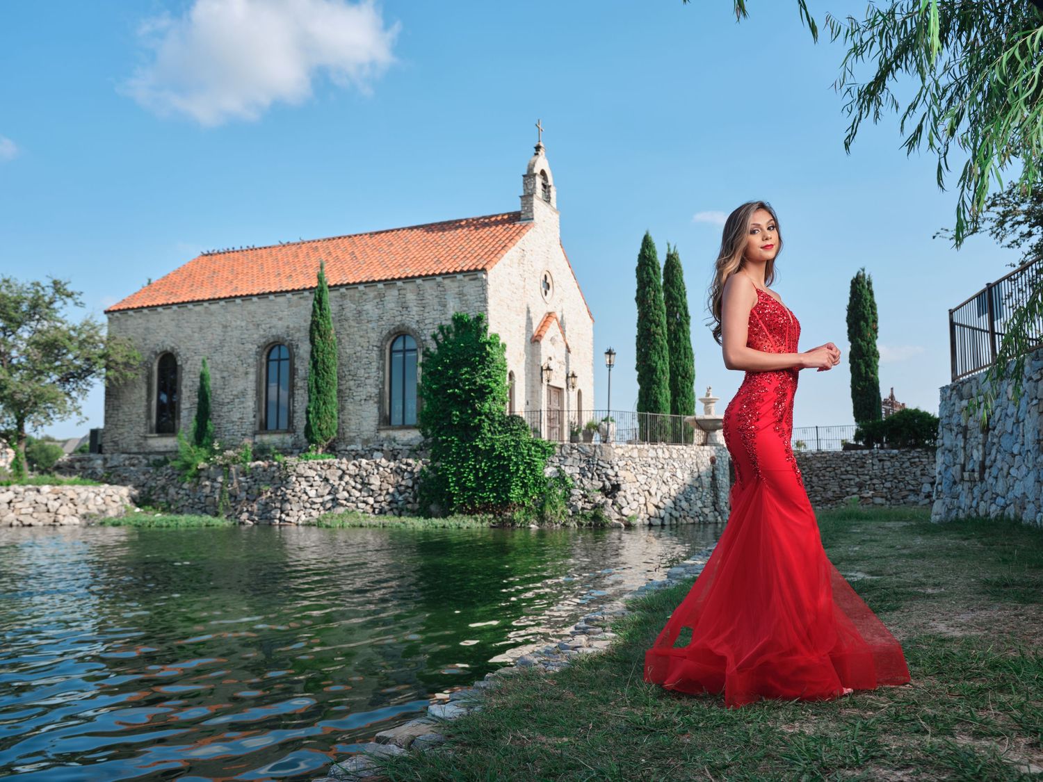 Elegant woman in red mermaid gown posing beside scenic lakeside chapel with cypress trees.