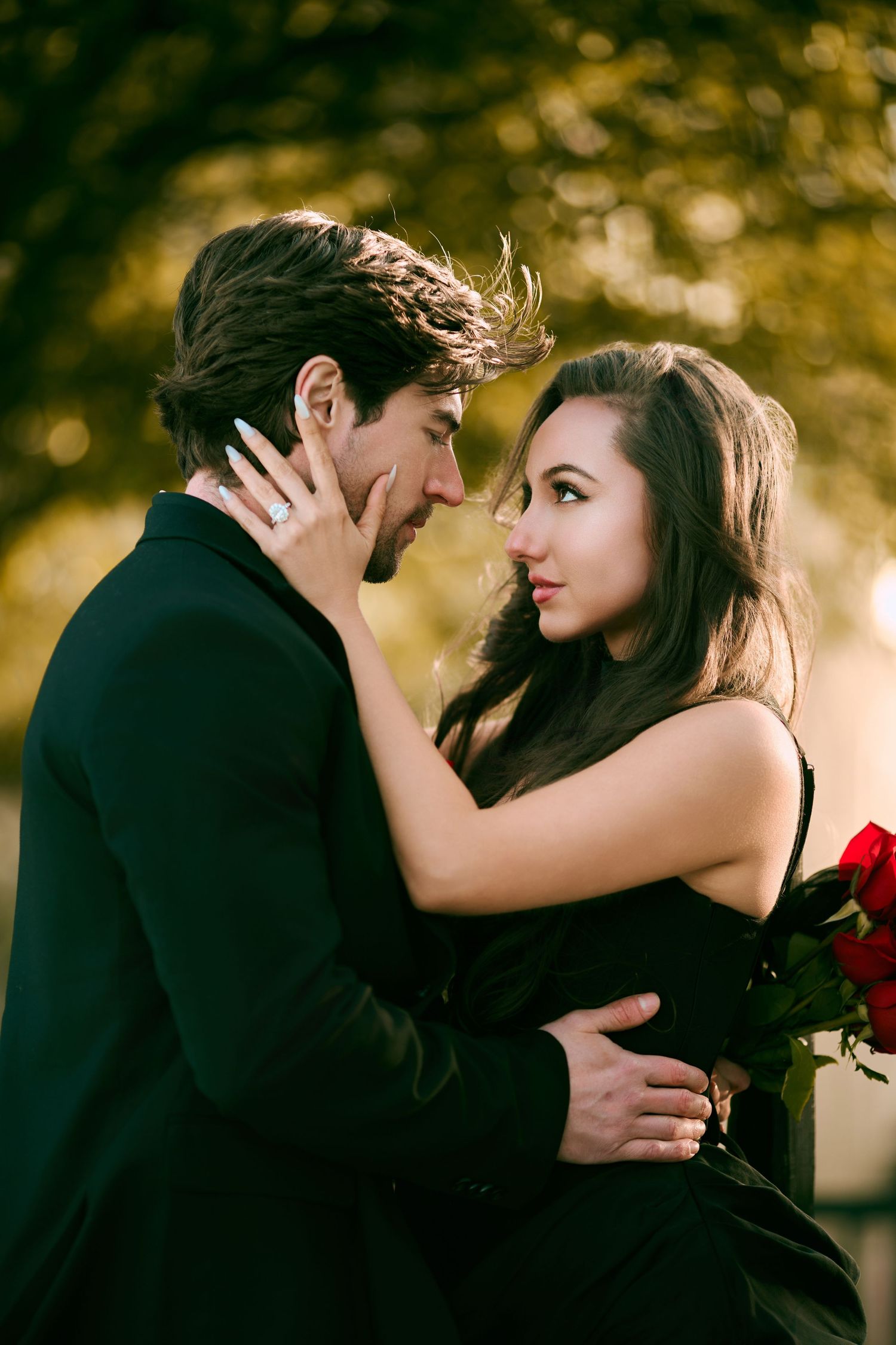 Romantic couple in black outfits embracing outdoors, woman holding red roses, sharing an intimate moment with golden bokeh background.