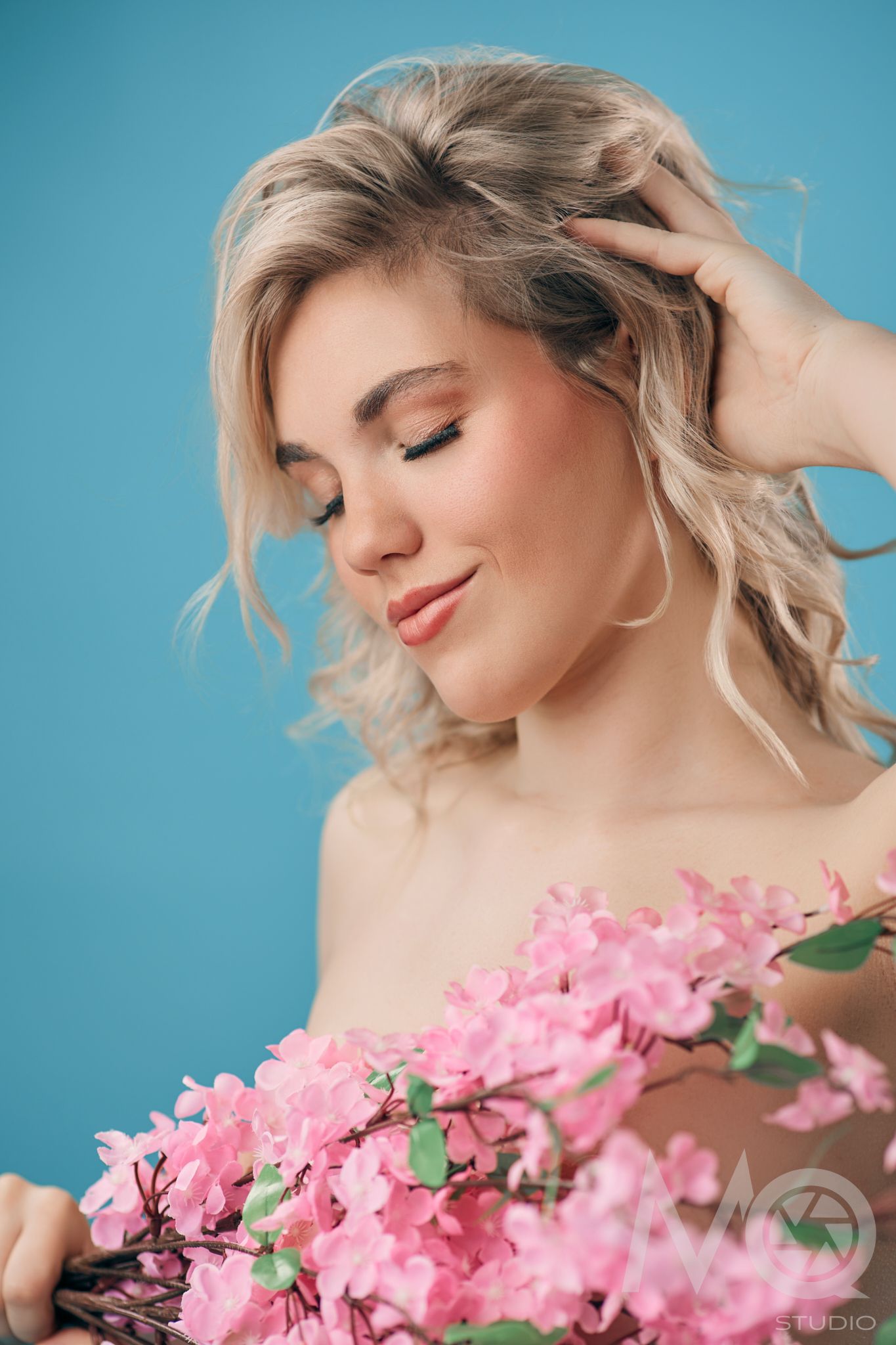Blonde woman with closed eyes holding pink spring flowers against a blue background.