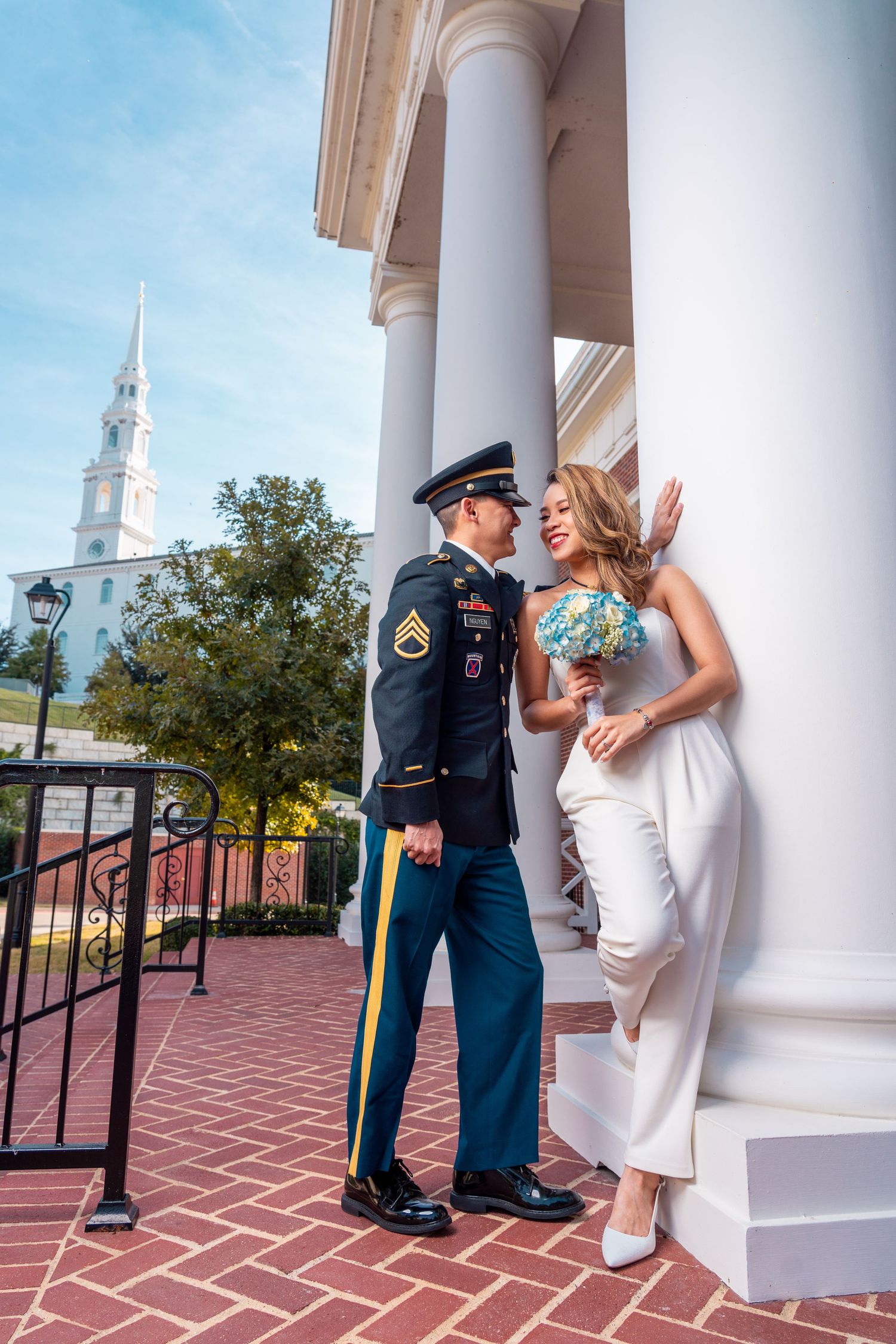 Military groom in dress uniform with bride in white holding floral bouquet by white columns.