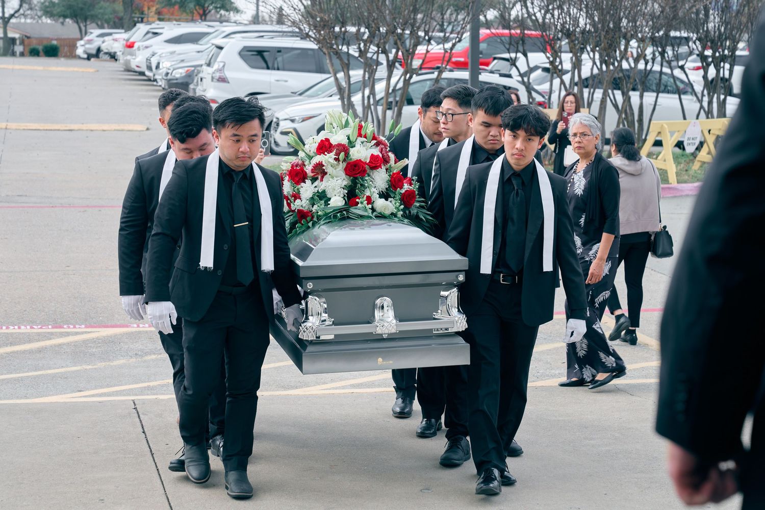 Pallbearers in black suits carrying a silver casket adorned with red and white flowers at a funeral.