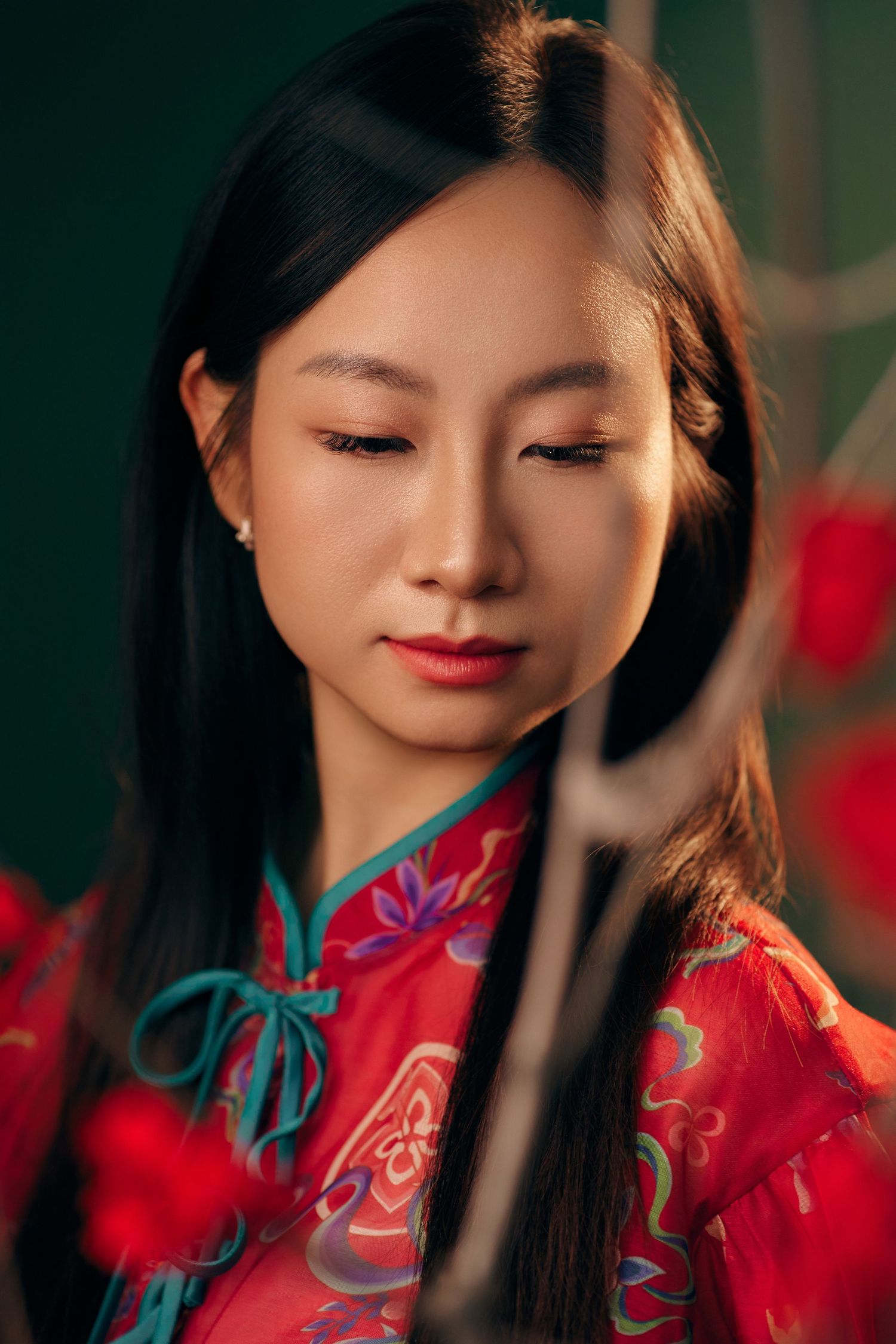 Young Asian woman in red floral traditional dress with eyes downcast on dark green background.
