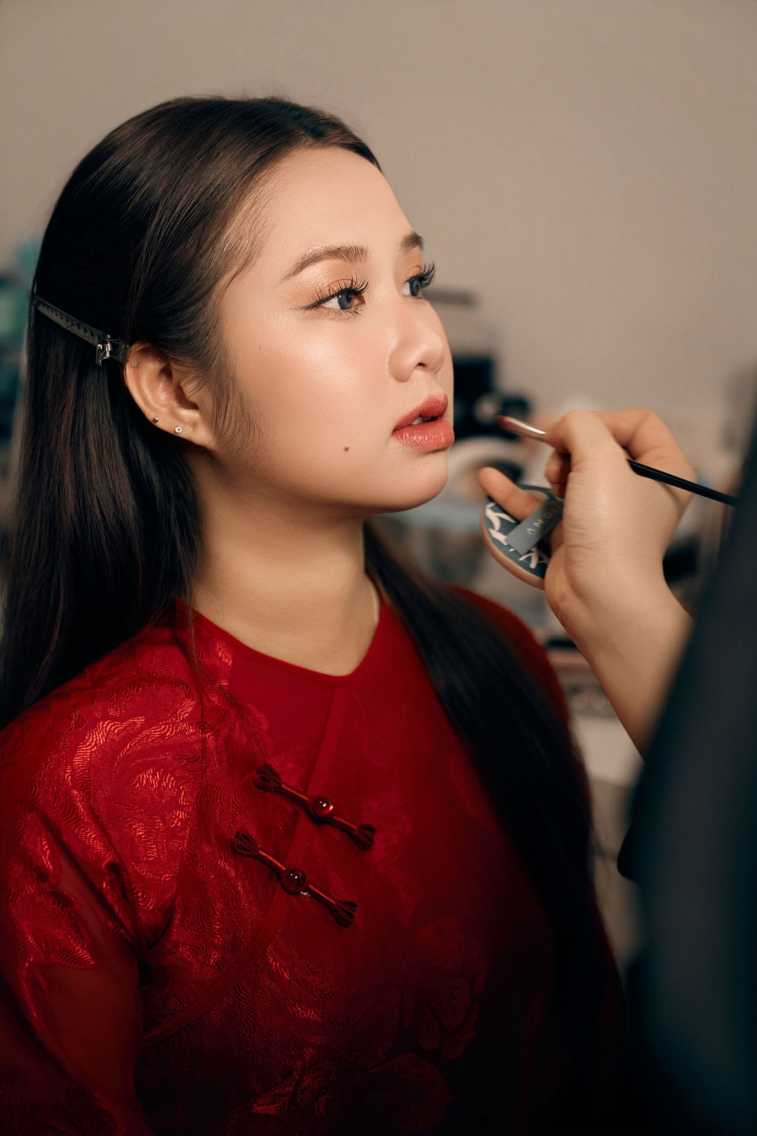 Young Asian bride in red traditional dress gets professional makeup applied before her wedding ceremony.