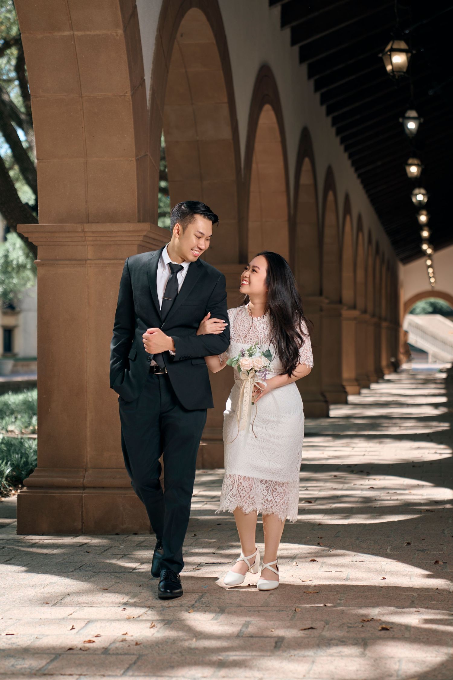 Asian couple in formal wear walking under arched colonnade, bride holding bouquet in white lace dress.