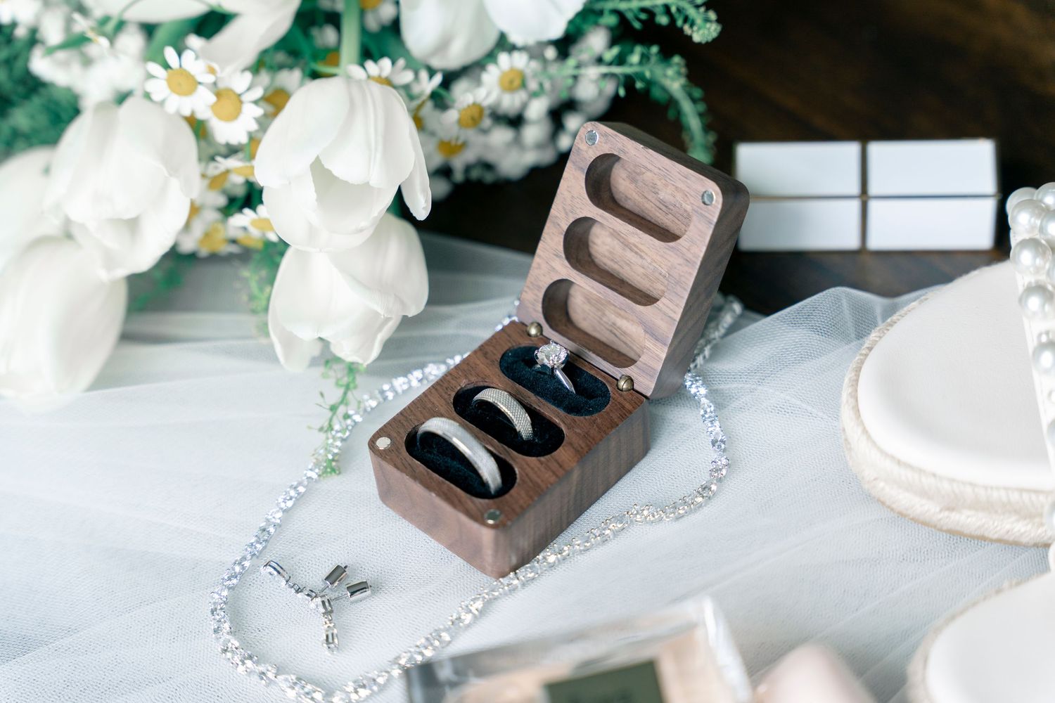 Elegant wooden wedding ring box displaying engagement and wedding bands with floral arrangements in the background.