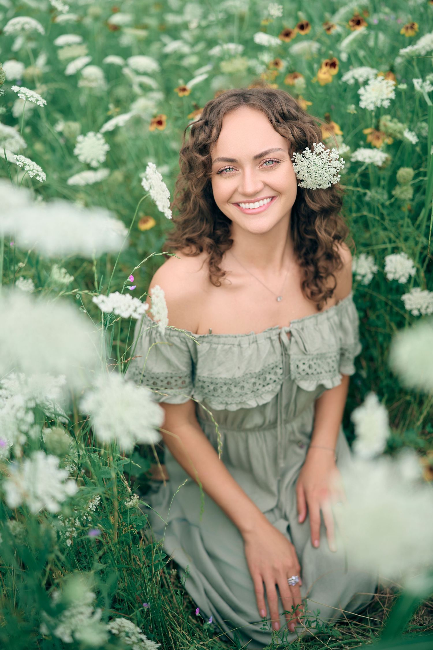 Smiling young woman in off-shoulder sage dress sitting among white wildflowers in a lush green field.