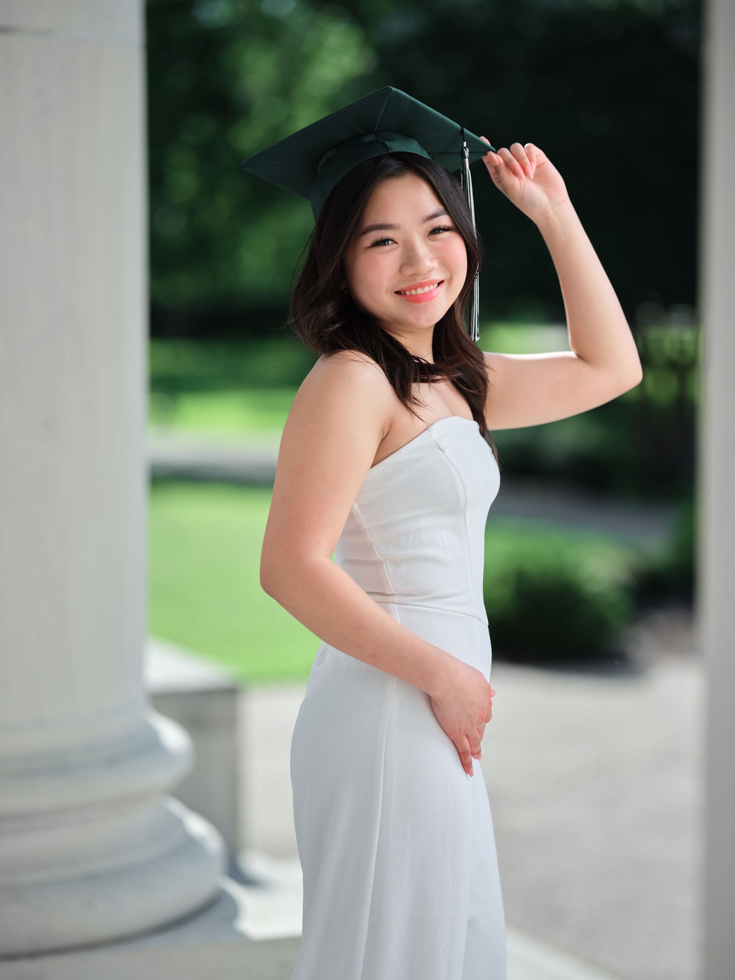 Smiling young woman in white dress holding a green graduation cap outdoors near columns.
