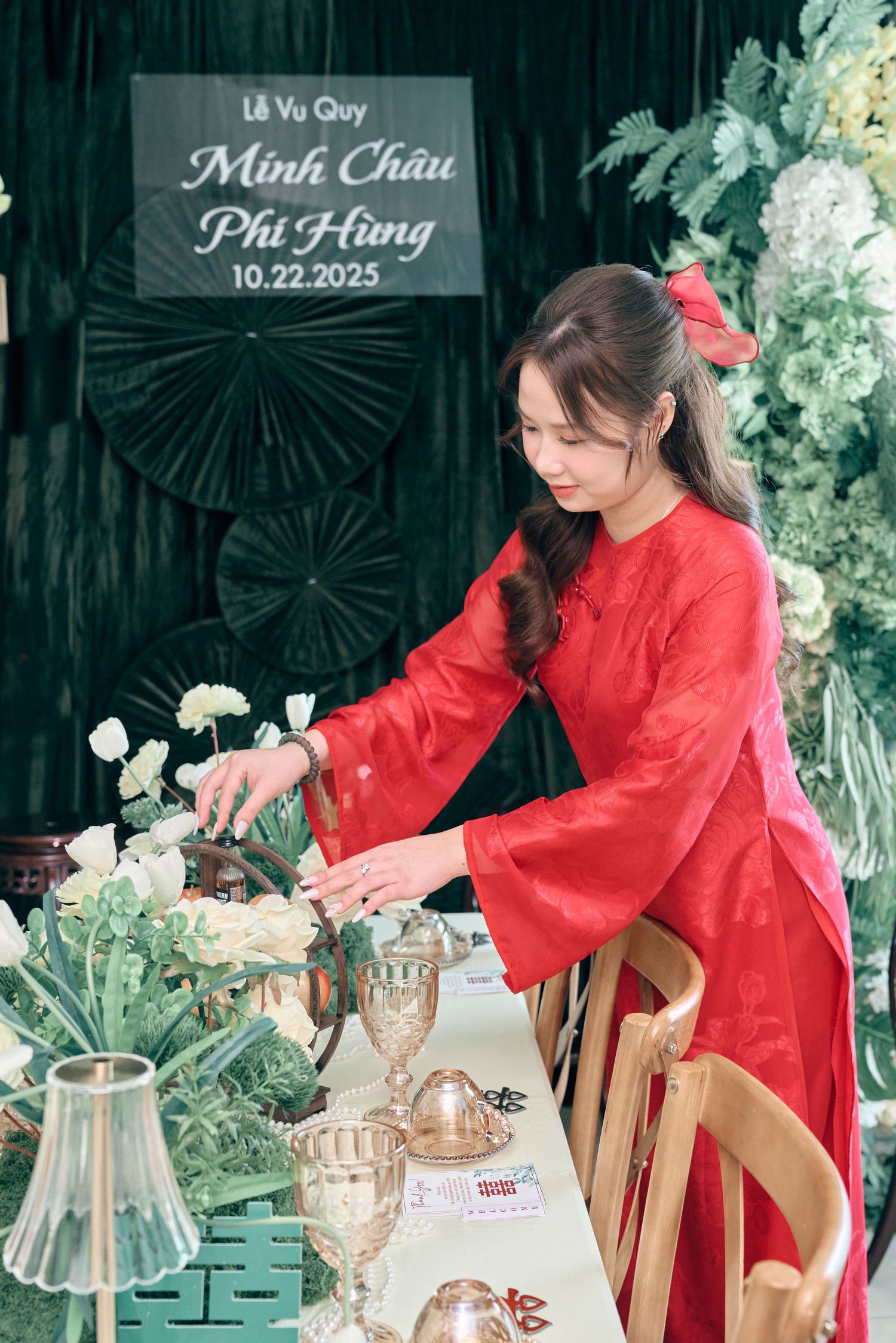 Woman in red ao dai arranges flowers at a Vietnamese wedding ceremony table dated 10.22.2025.