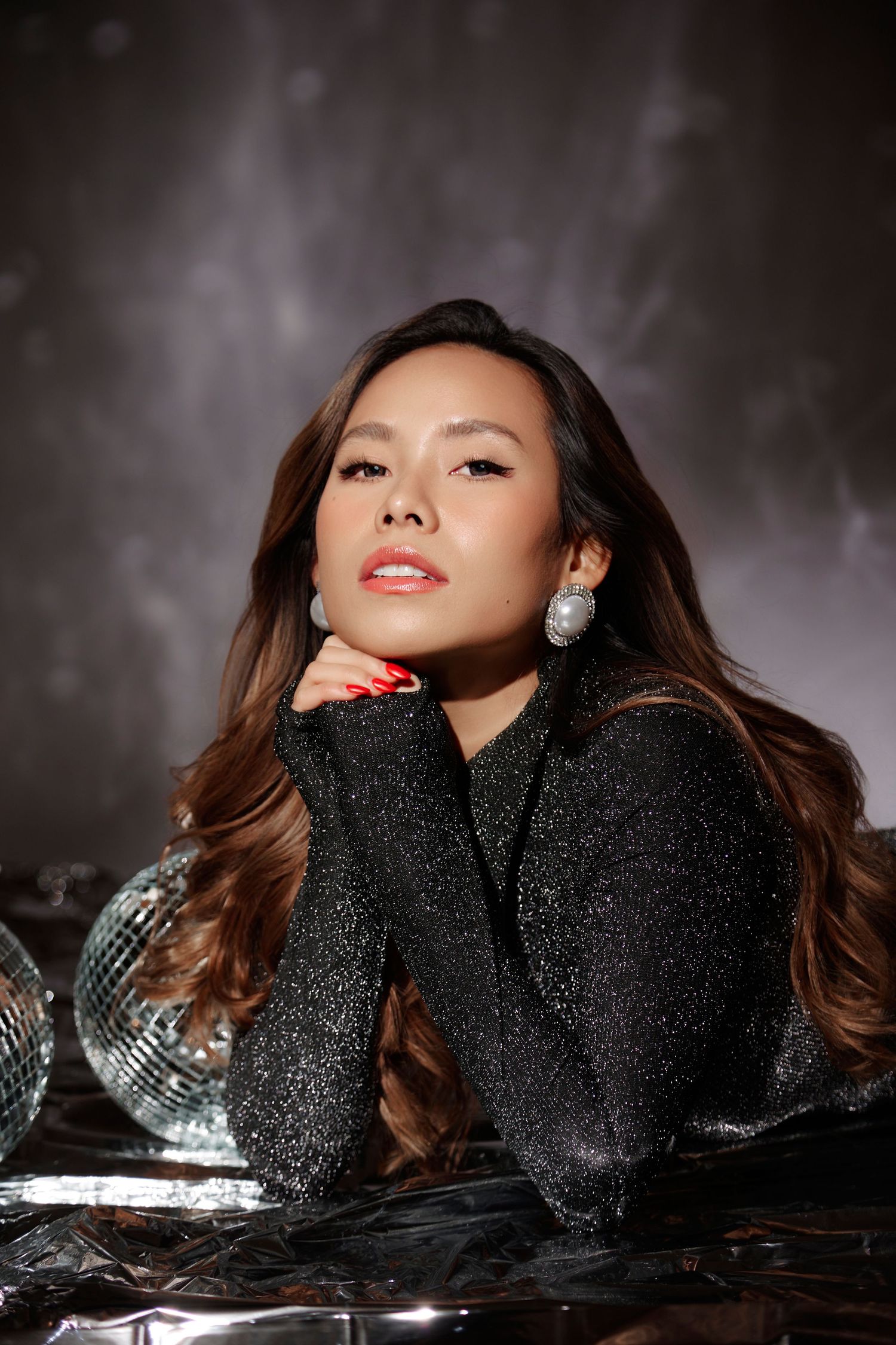 Stylish Asian woman in black sparkle turtleneck with pearl earrings posing with disco balls in moody studio.