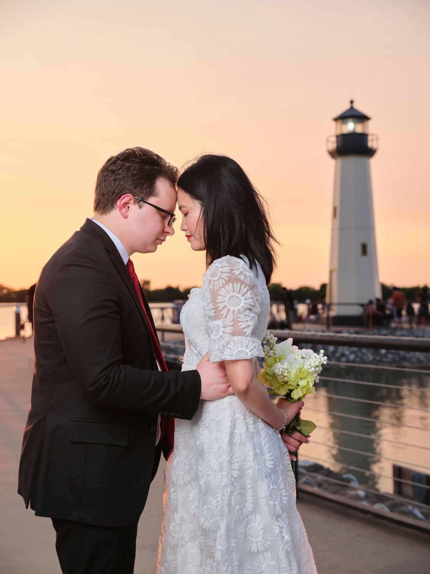 Couple sharing romantic moment at sunset near lighthouse, bride in lace dress holding white bouquet.