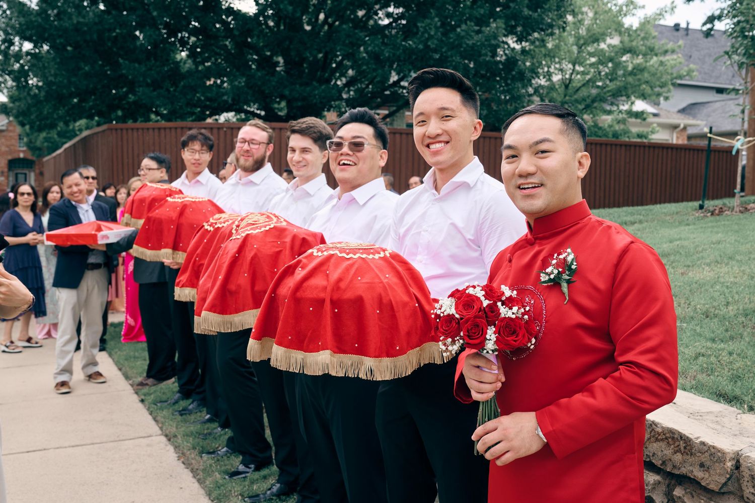 Groomsmen in red traditional Chinese wedding attire holding decorative red trays outdoors during a wedding ceremony.
