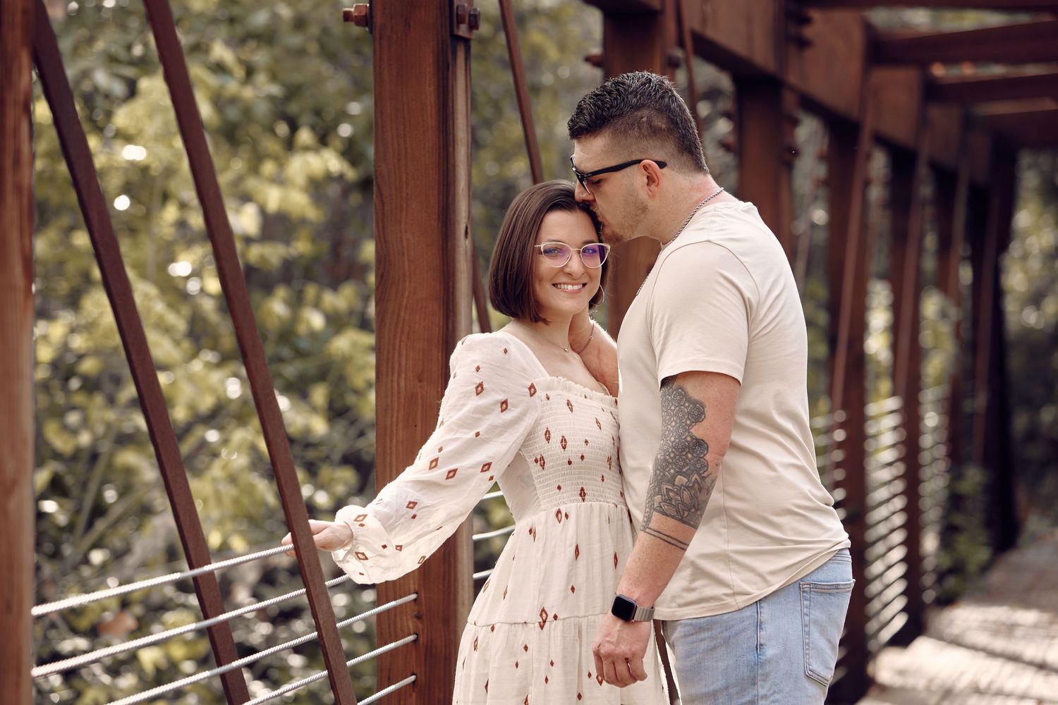 Smiling couple holding hands on a wooden bridge surrounded by lush green trees.