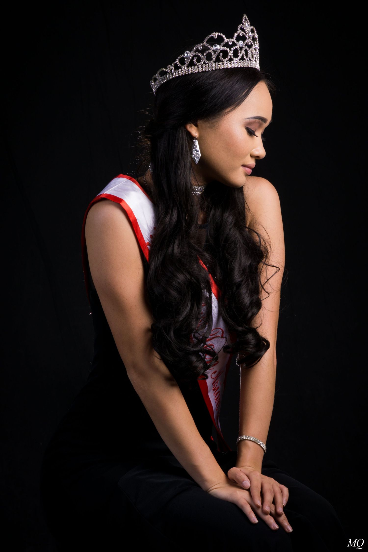Elegant woman wearing a tiara and pageant sash with long wavy hair posing on a dark background.