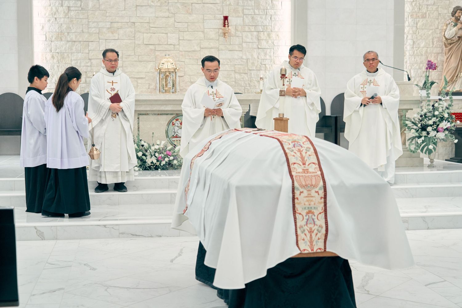 Catholic priests in white vestments performing funeral rites over a draped casket at a church altar.