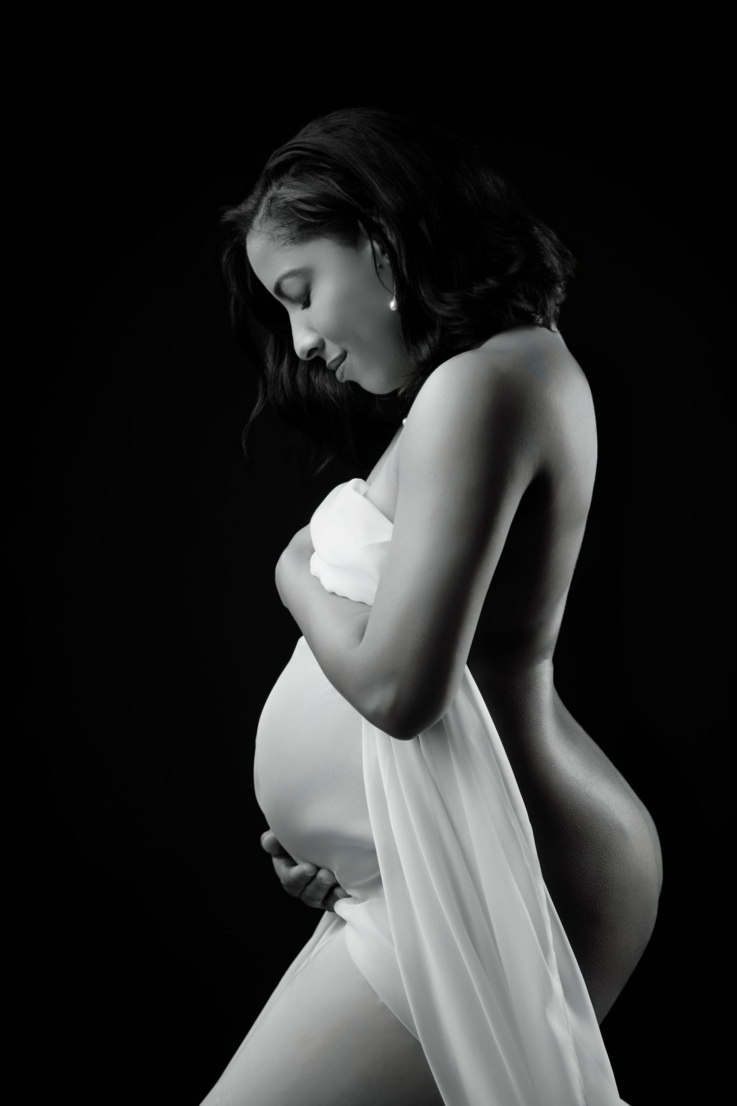 Elegant black and white portrait of pregnant woman in white draped fabric cradling her belly.