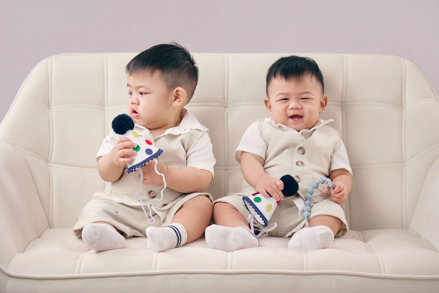Two Asian baby twins in matching beige outfits sitting on a white sofa, holding toy microphones.