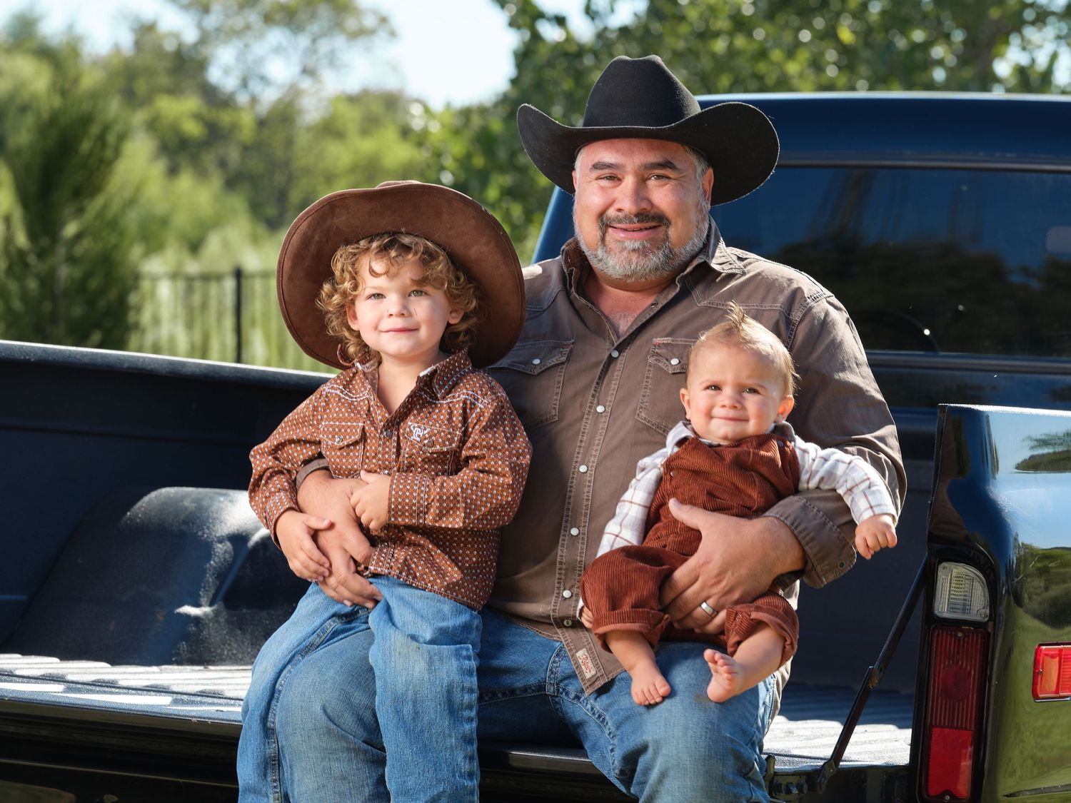 A father in a cowboy hat sits on a truck bed smiling with his two young sons in matching western outfits.