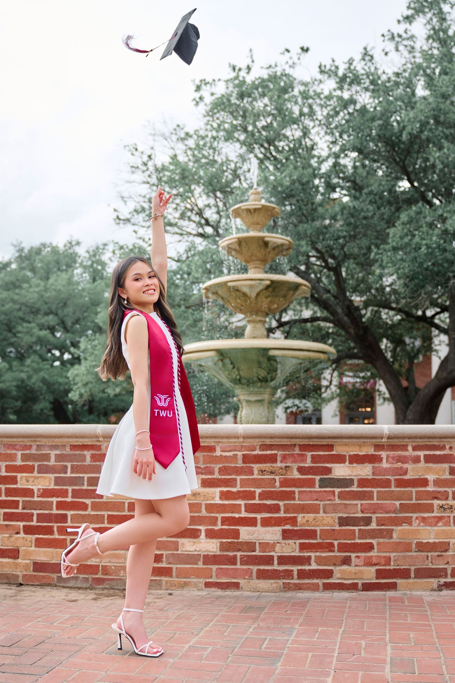 Graduate in white dress tosses cap near a brick fountain wearing a red stole.
