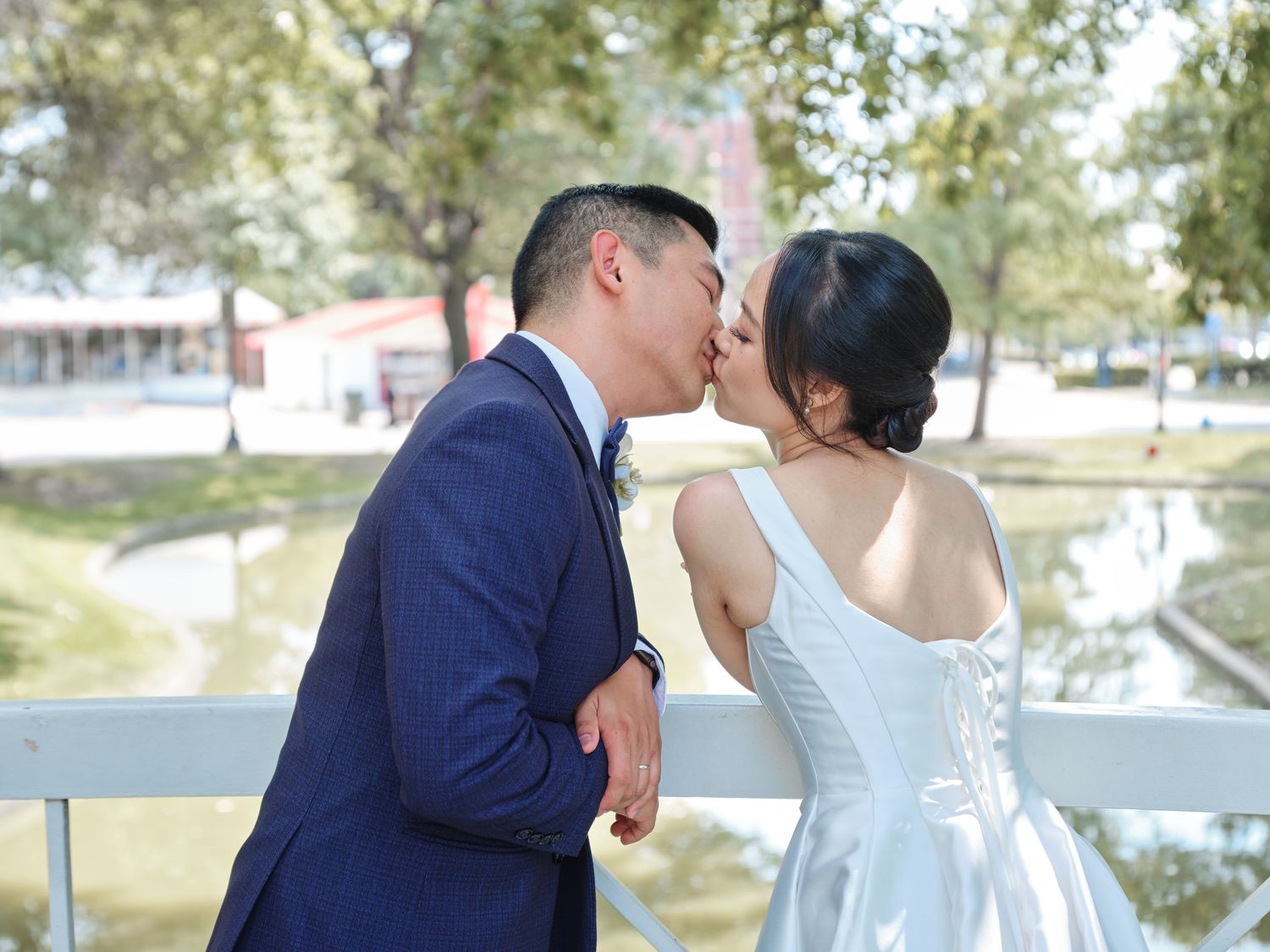 Asian newlywed couple sharing a kiss on white bridge overlooking serene pond in outdoor setting.