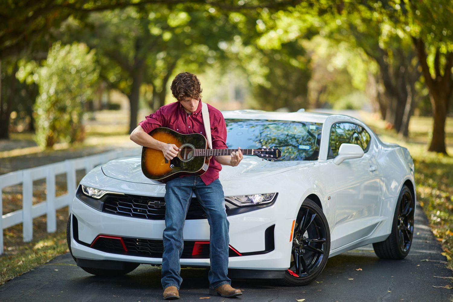Young man playing acoustic guitar leaning against a white Chevrolet Camaro on a tree-lined driveway.