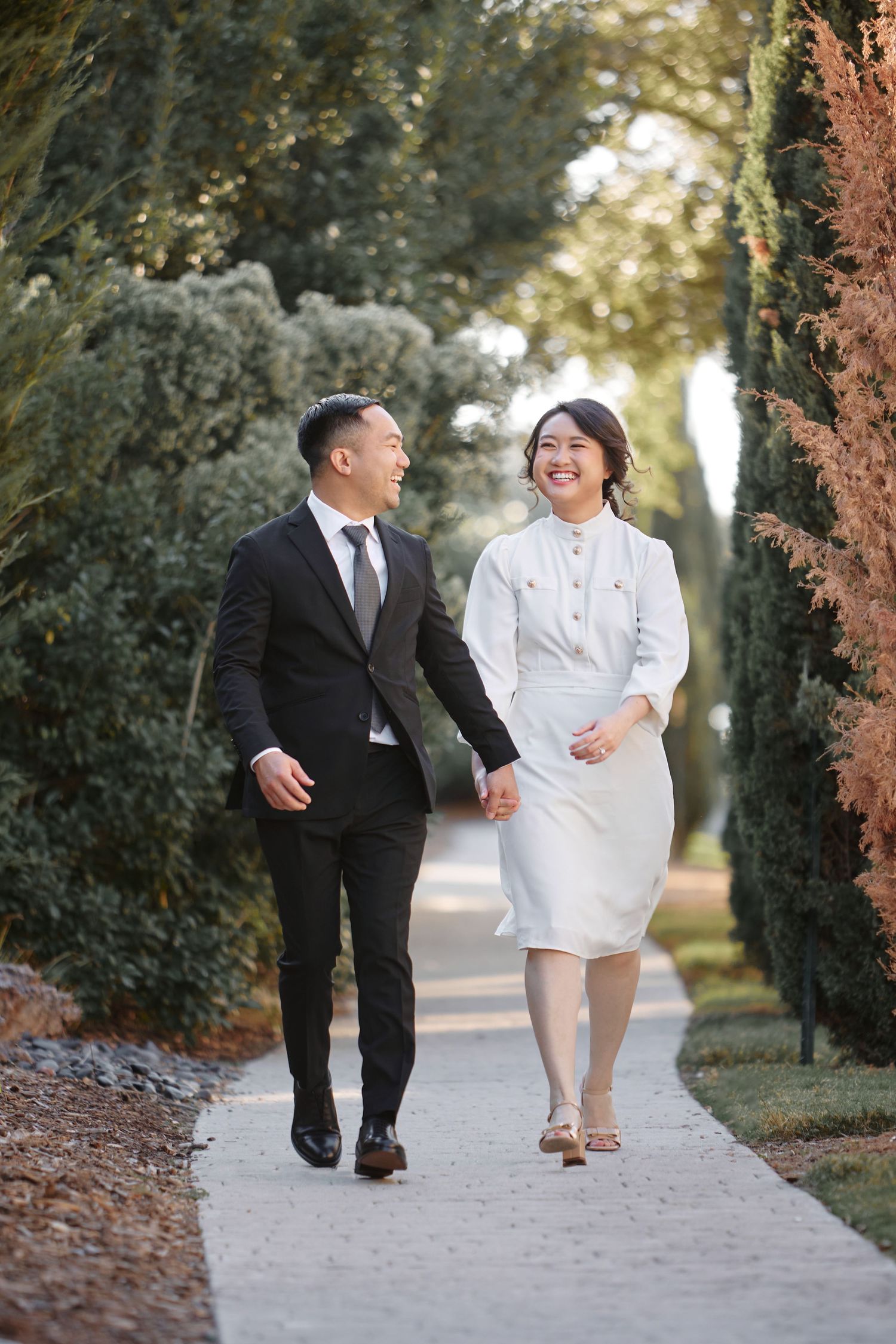 Happy Asian couple in formal attire walking hand in hand along a tree-lined path.