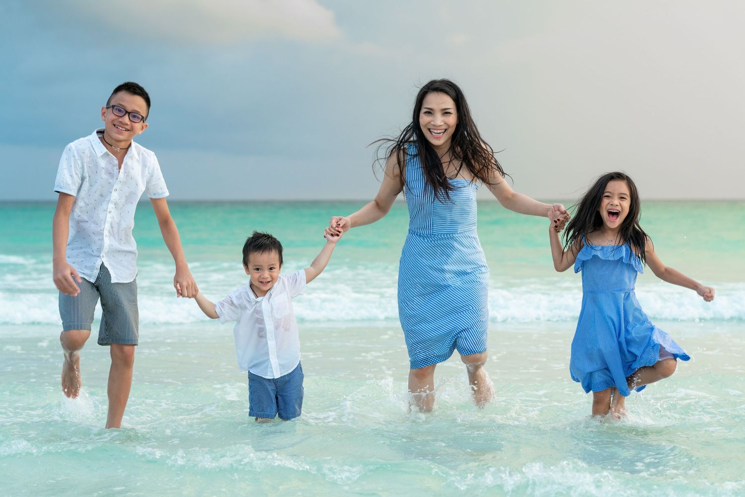 Happy family of four splashing in ocean waves on a sunny beach, wearing coordinated blue and white outfits.