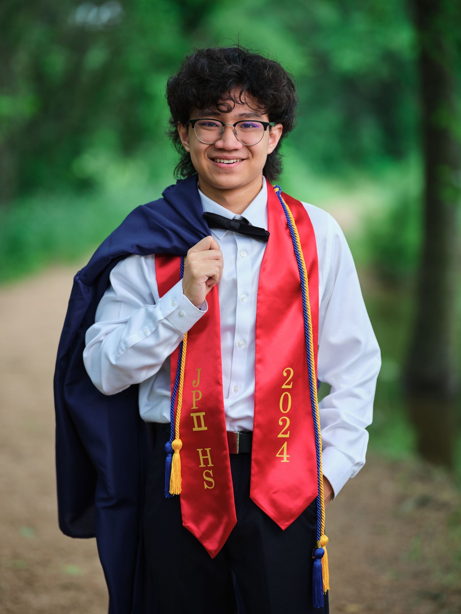 Smiling young man in graduation gown displays red 2024 honor stole on a path.