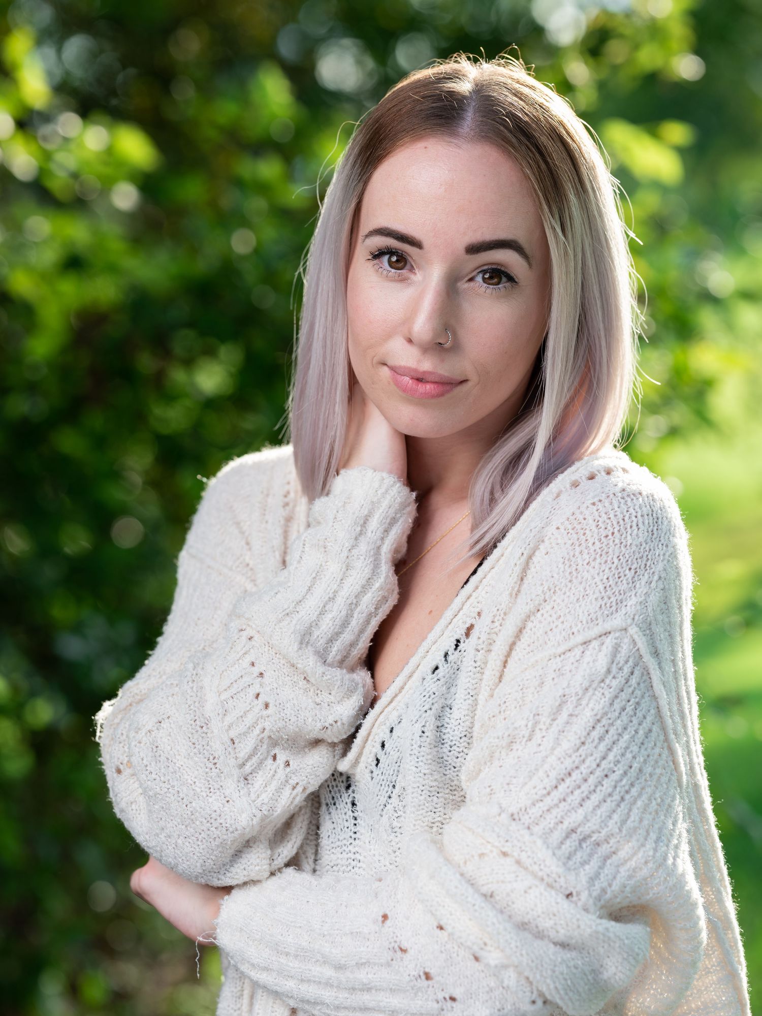 Young blonde woman in a cream knit sweater poses outdoors with green bokeh background.