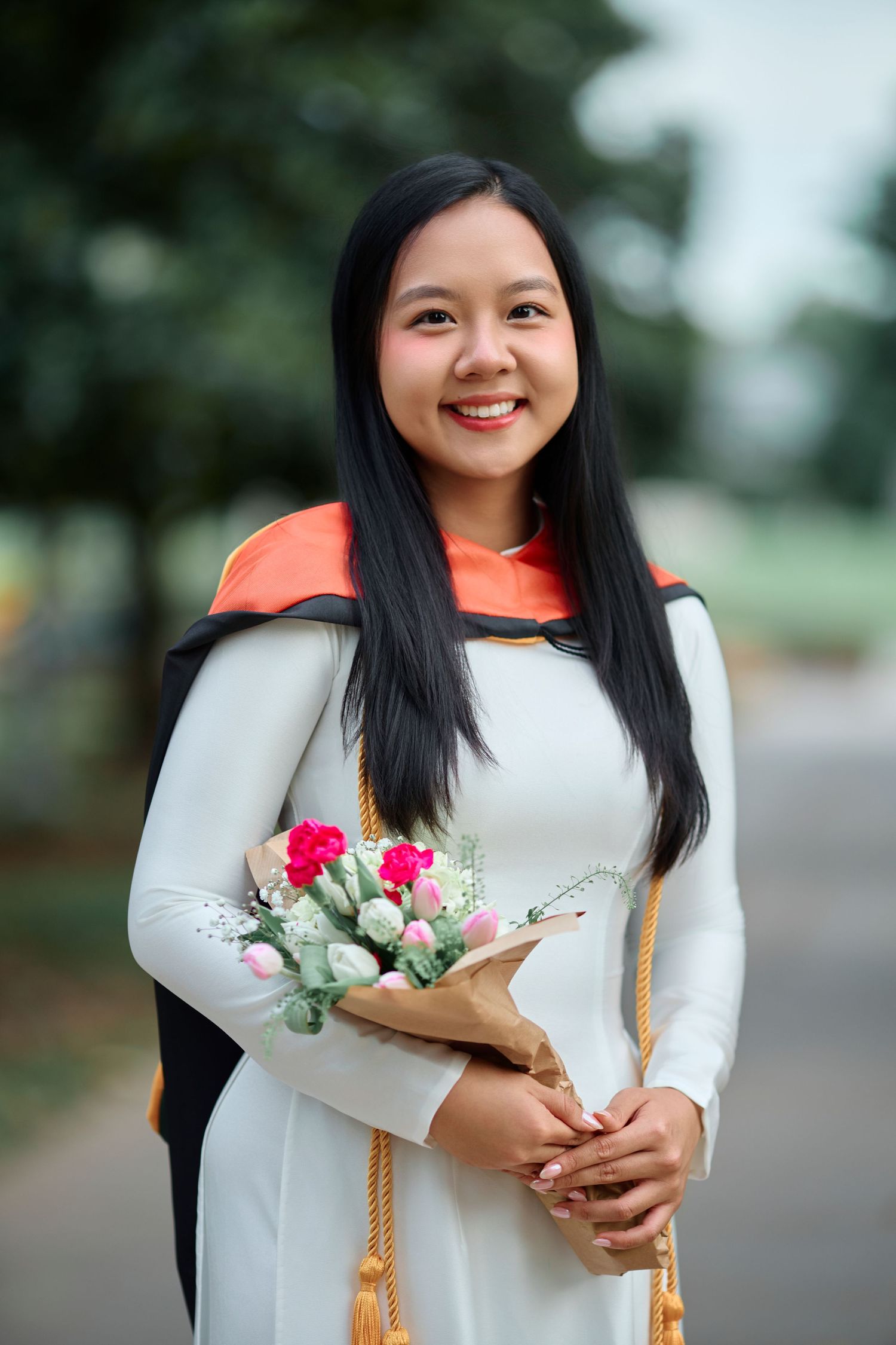 Smiling graduate in white dress holds flower bouquet with orange academic hood.
