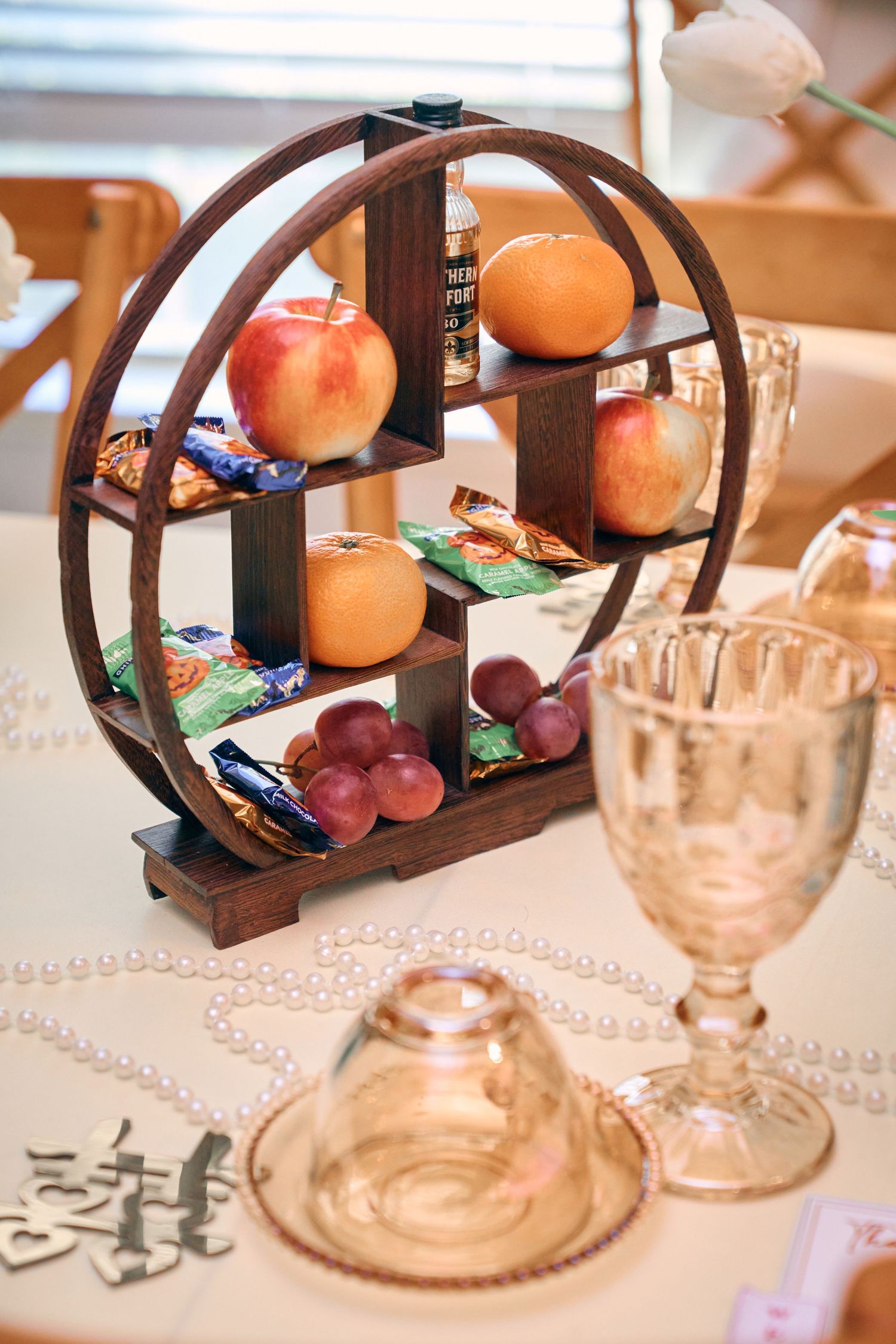 Wooden circular tiered display stand with apples, oranges, grapes, and snacks beside crystal glasses on a white tablecloth.
