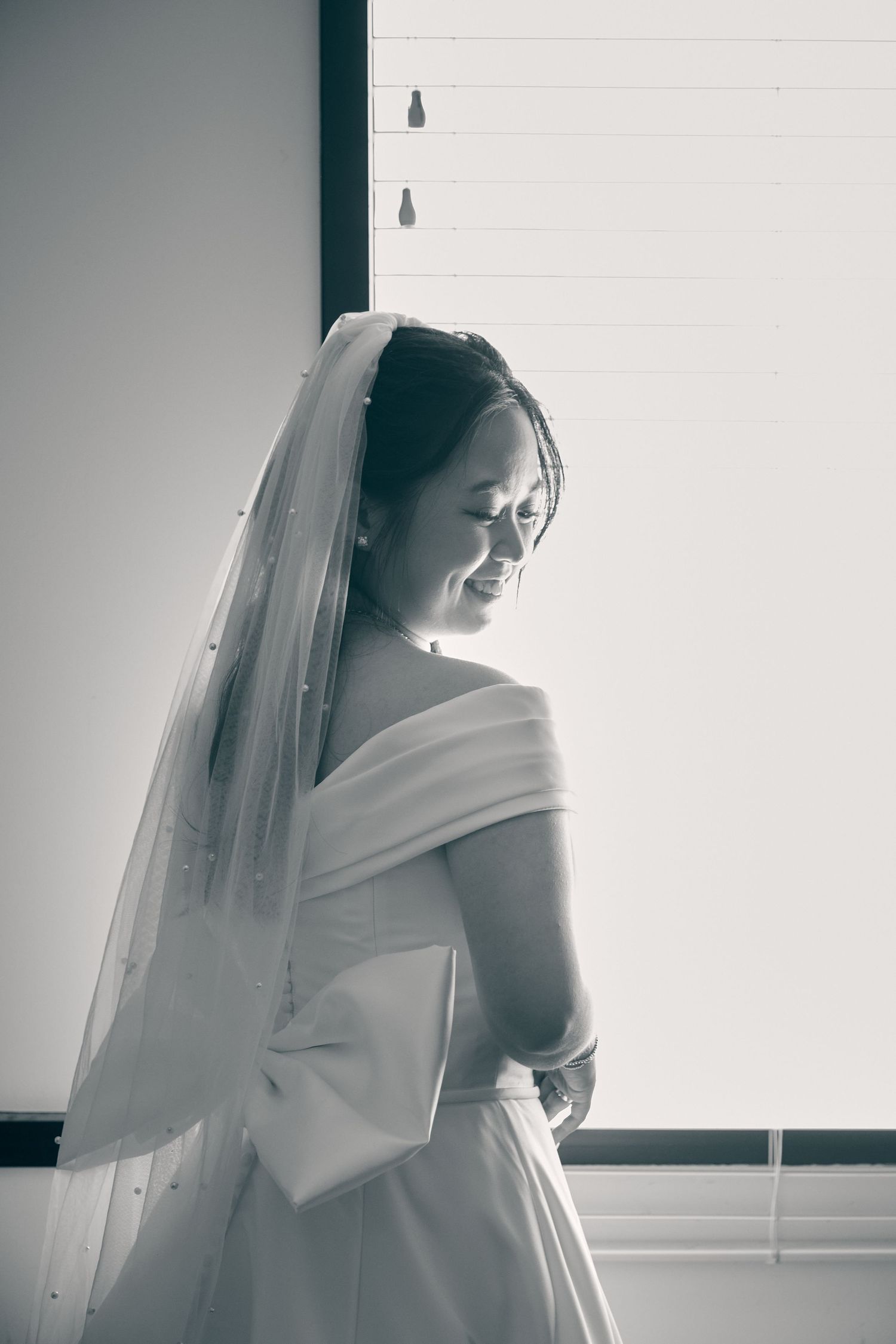 Elegant bride in white off-shoulder gown and veil smiling softly by a bright window, black and white.