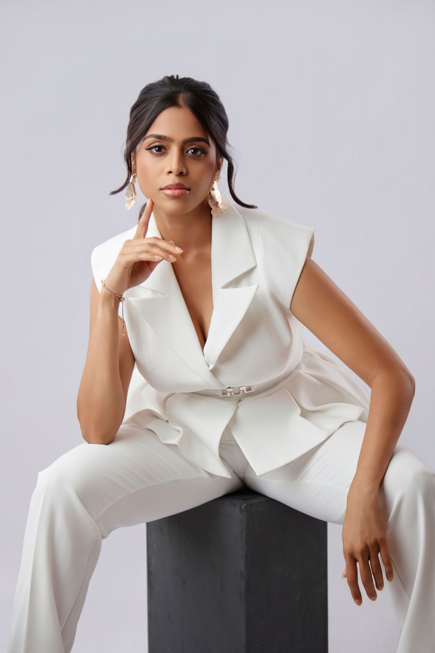 Elegant South Asian woman in white sleeveless blazer suit sits on dark cube in studio.