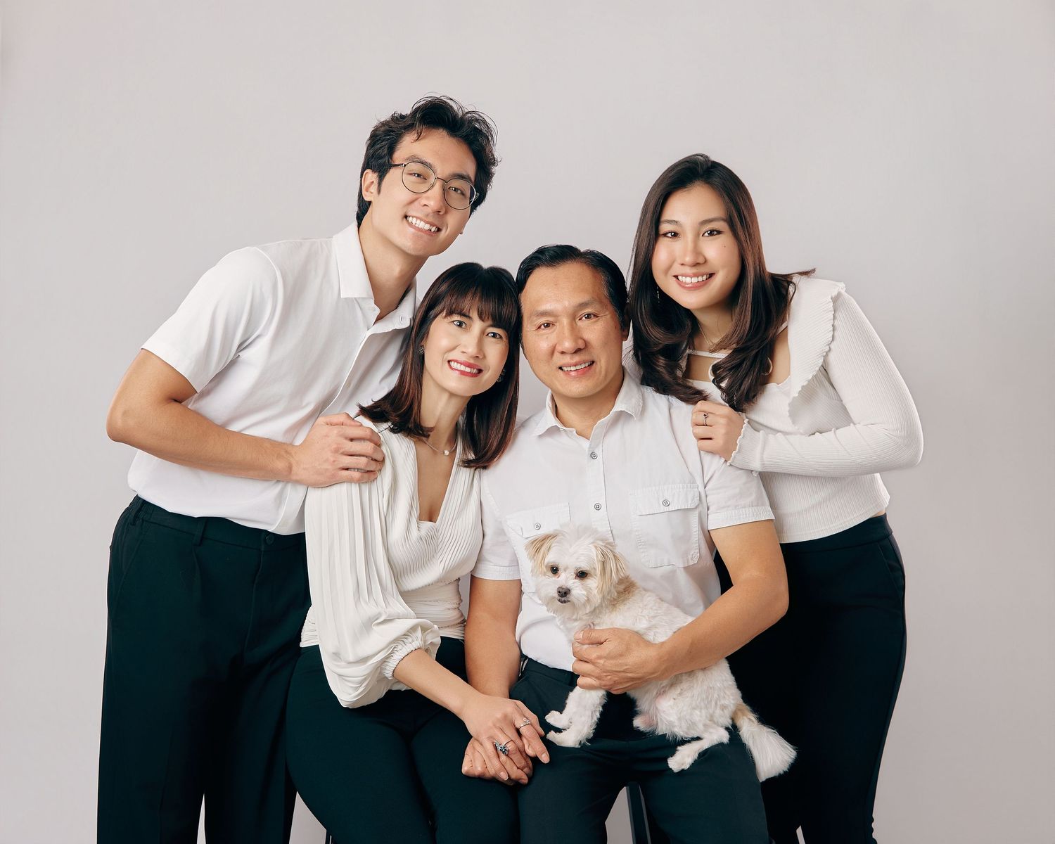 Asian family of four with a small white dog posing together in matching white tops for studio portrait.