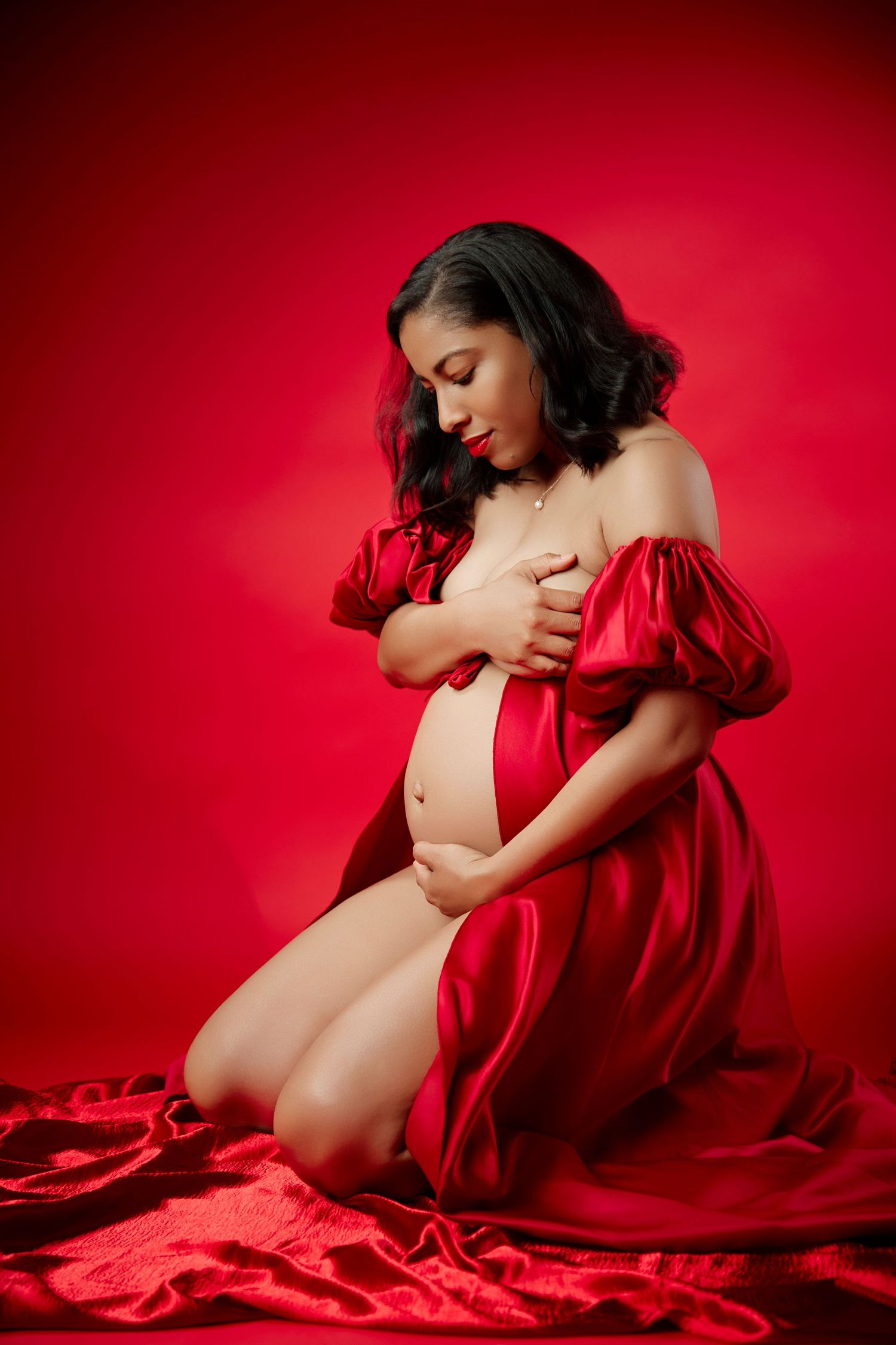 Pregnant woman kneeling on red satin fabric in elegant red off-shoulder dress against red background.
