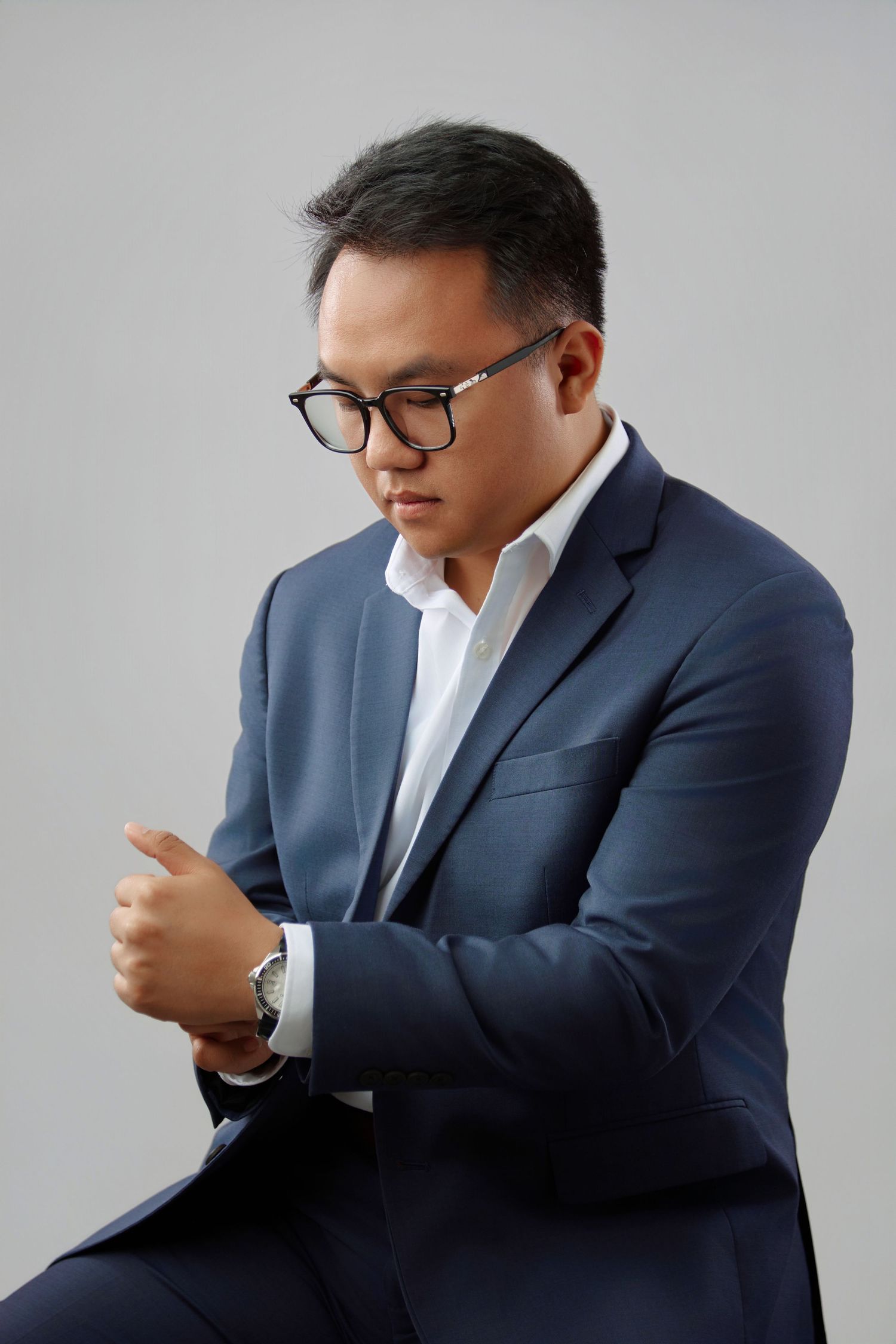 Young Asian man in navy blue suit adjusting sleeve while looking down in studio.