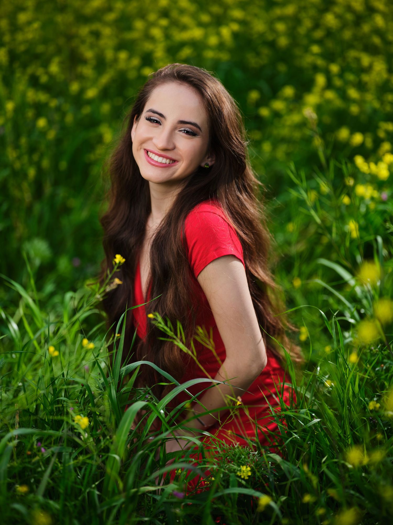 Happy young woman with long brown hair sitting in a vibrant yellow wildflower field wearing a red dress.