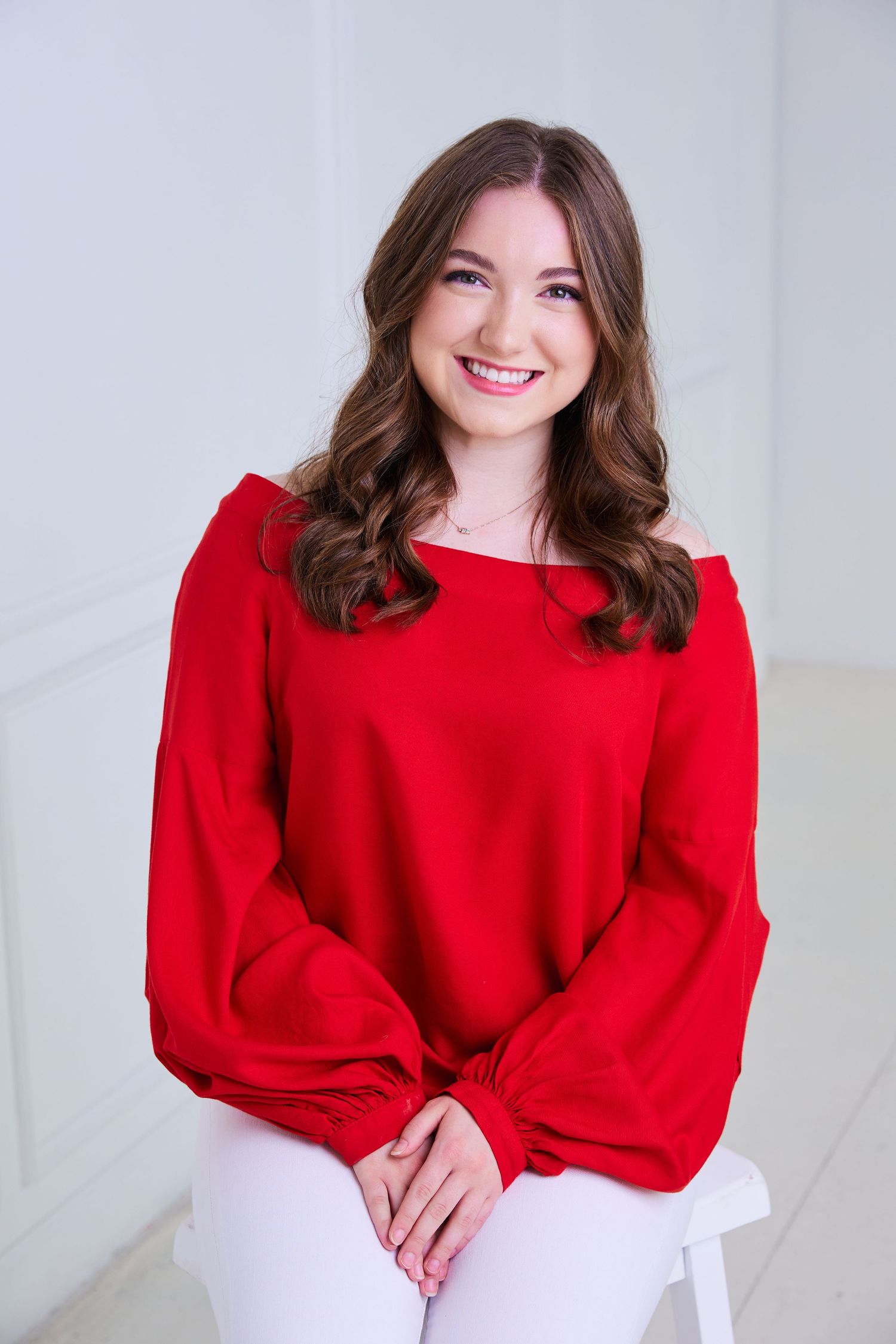 Cheerful young brunette woman in a red off-shoulder top sitting and smiling in a bright white studio.