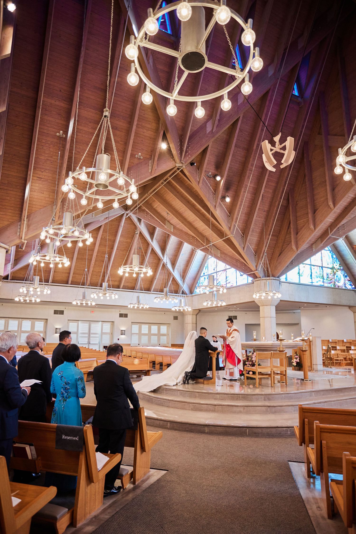 Wide angle view of church wedding ceremony with vaulted wooden ceiling and decorative chandeliers overhead.
