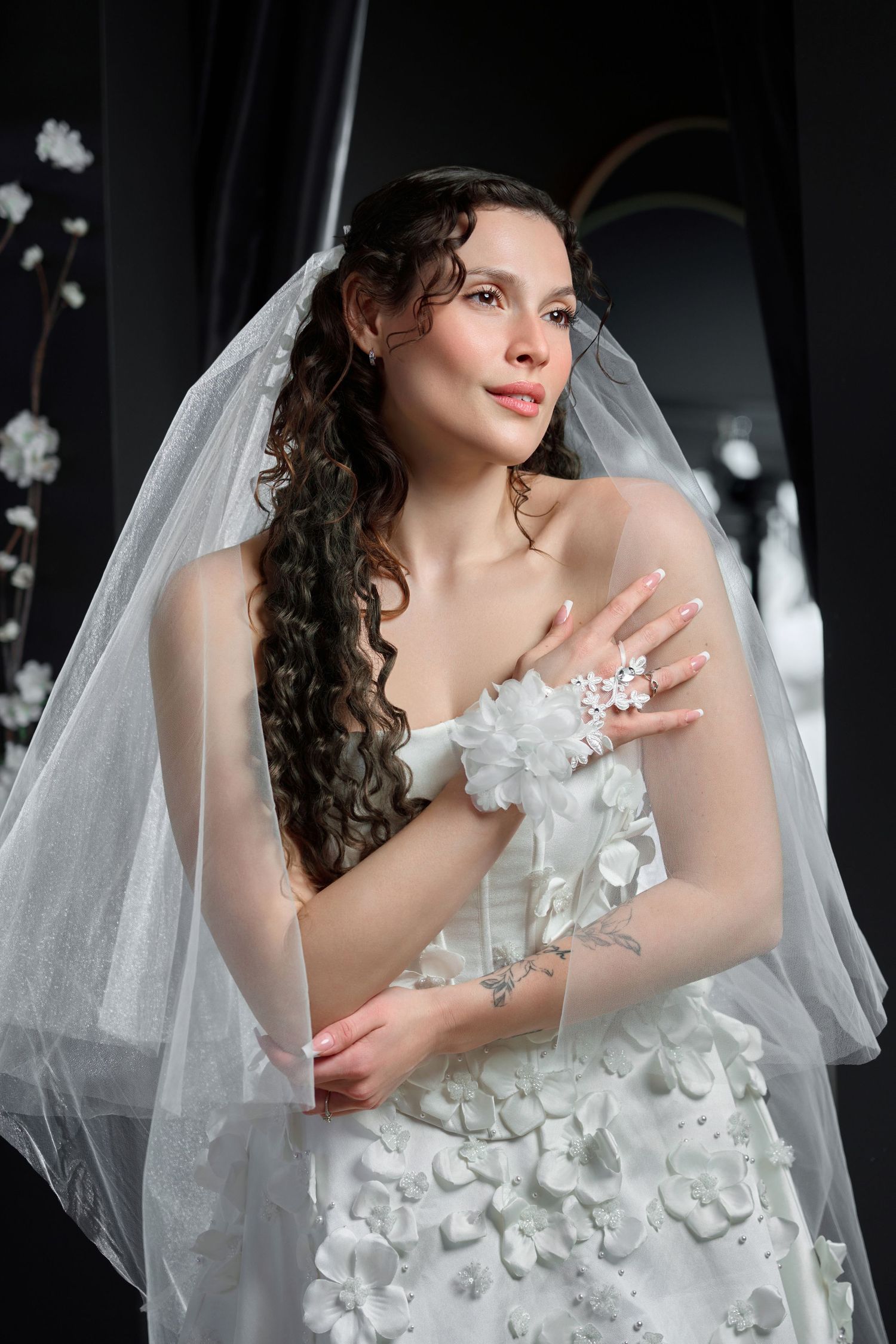 Beautiful bride in white floral wedding dress with veil and wrist corsage posing indoors.