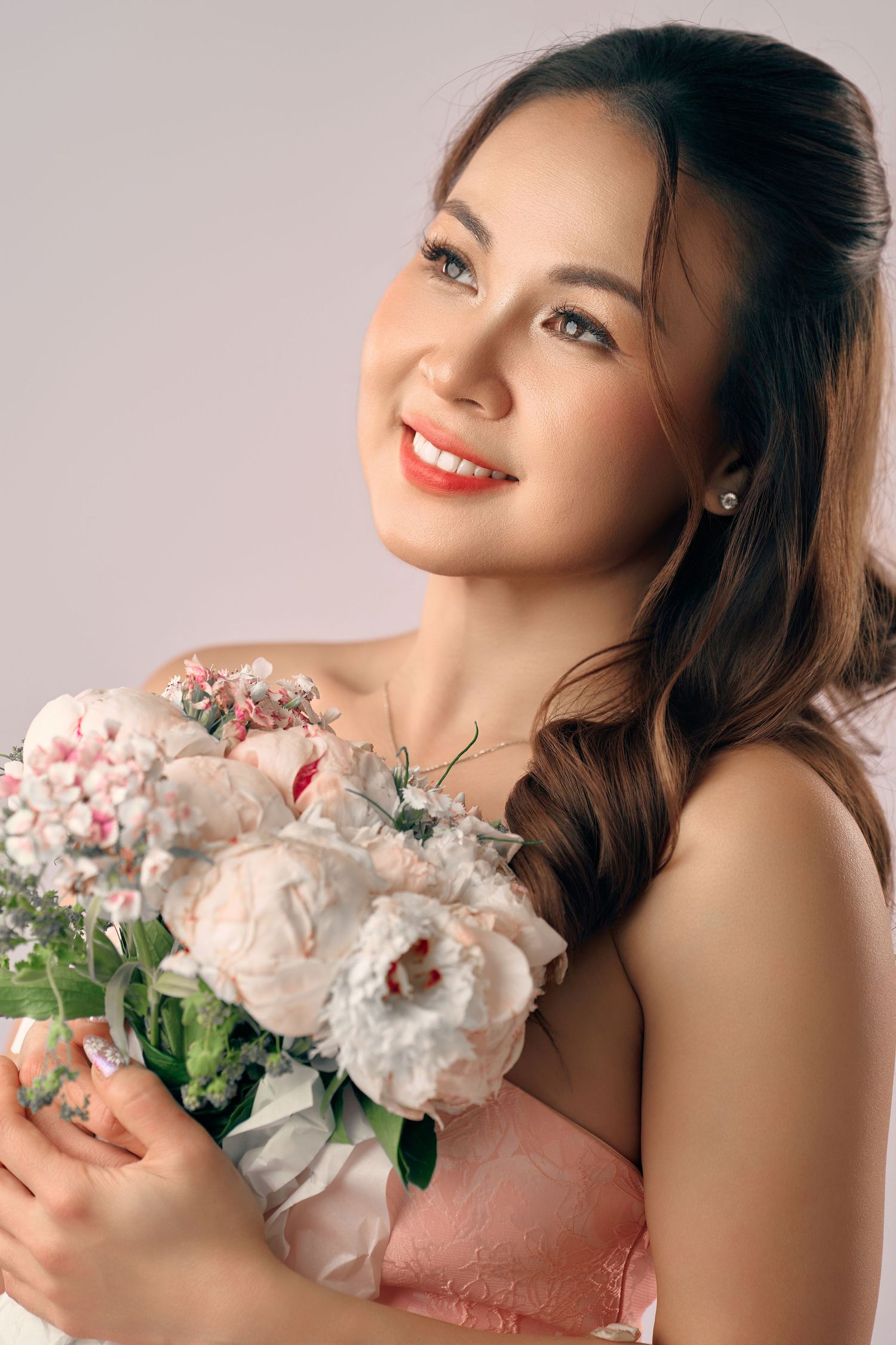 Smiling young woman holding a bouquet of white and pink peonies, wearing a pink dress, with elegant makeup.
