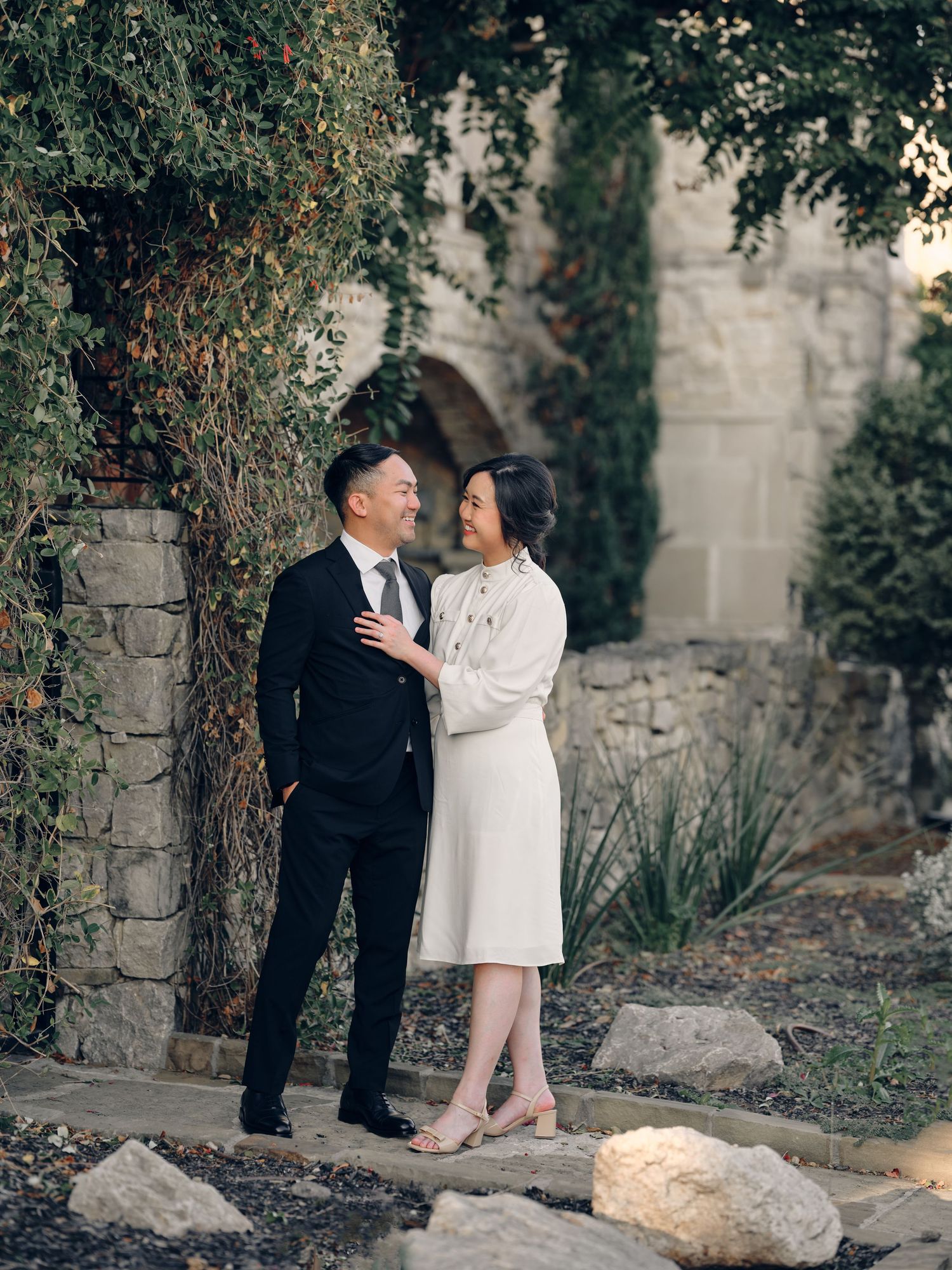 Couple in formal attire sharing a romantic moment by a stone wall with lush greenery backdrop.