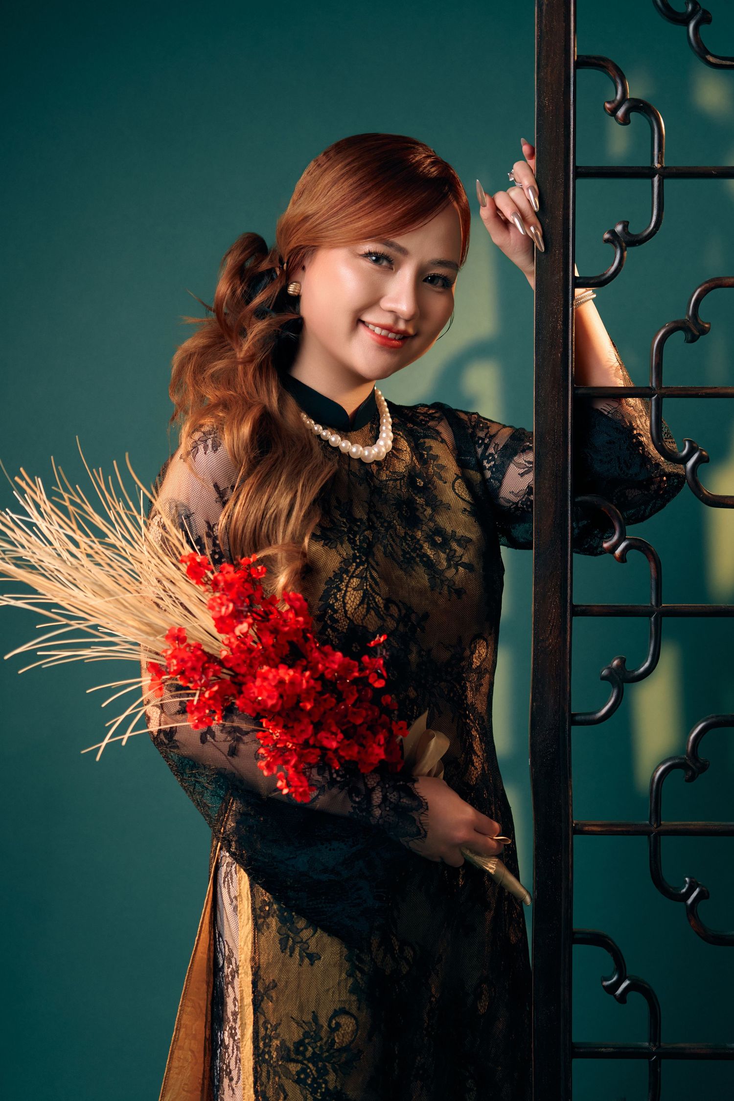 Smiling woman in black lace ao dai holding red flowers beside ornate metal gate.