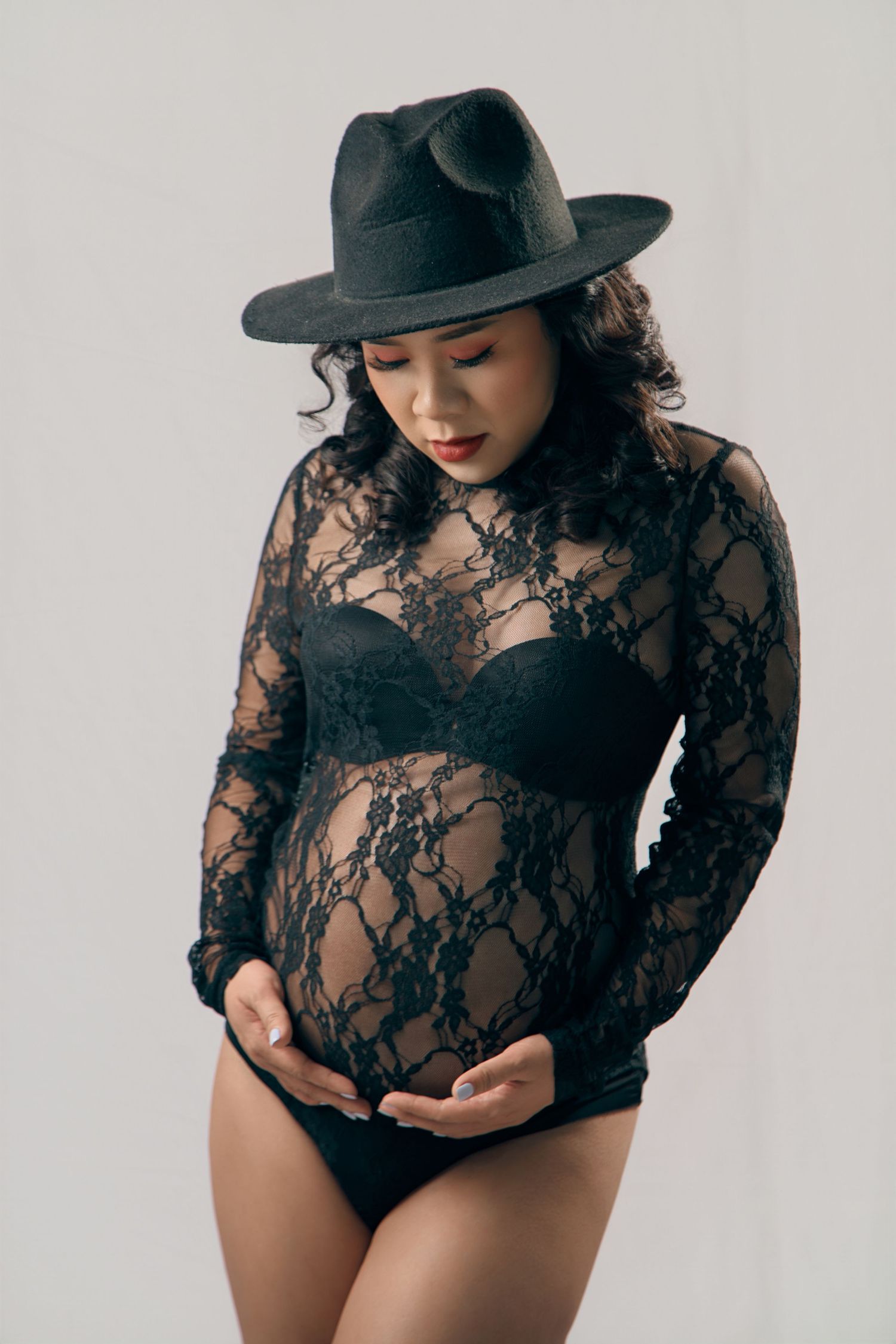 Pregnant woman in black lace bodysuit and fedora hat cradling her baby bump in a studio setting.