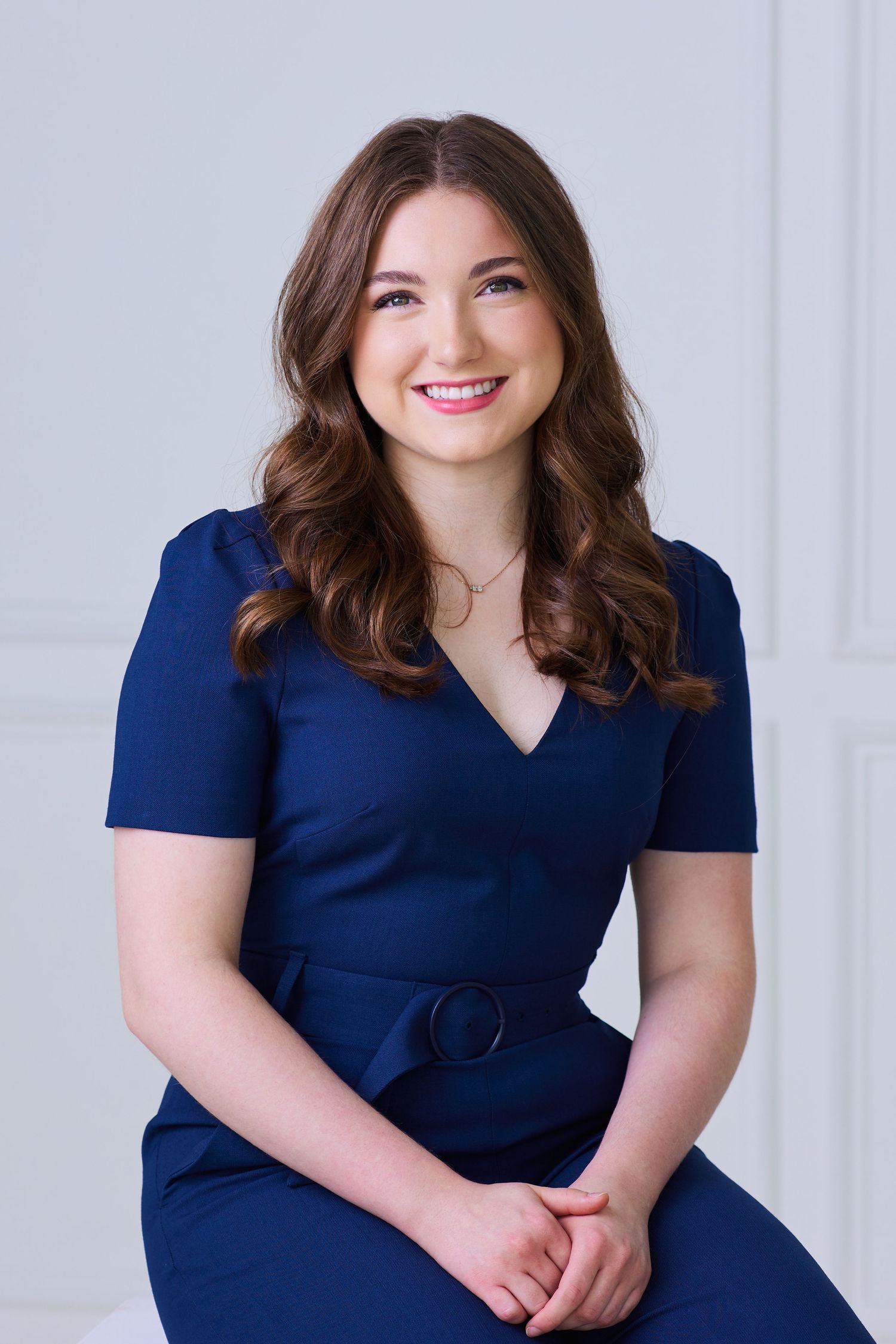 Smiling young woman in navy blue belted dress poses in bright white room.
