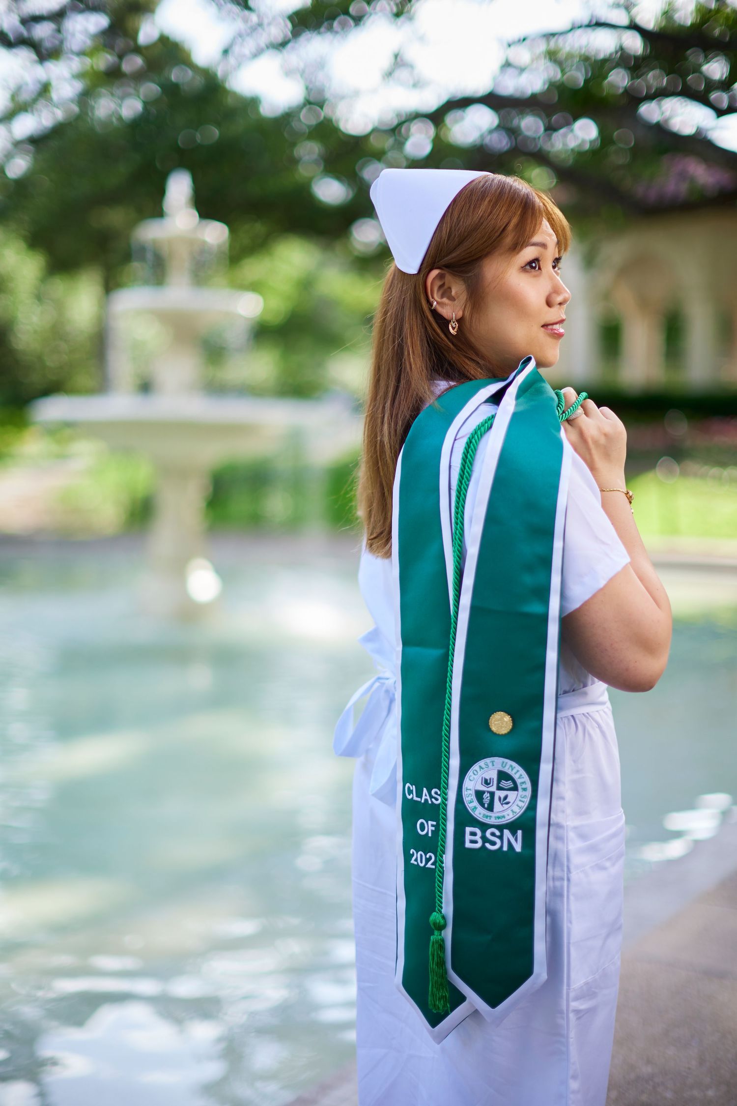 Nursing graduate in white uniform displays green BSN Class of 2022 graduation stole near fountain.