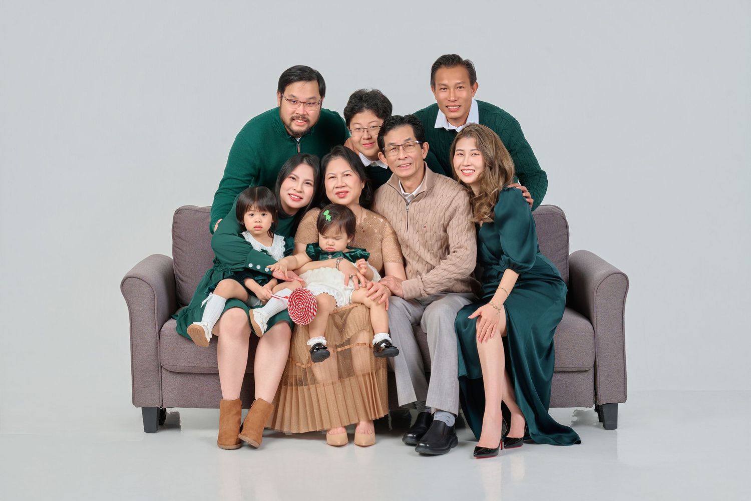 Multi-generational Asian family portrait with adults and children gathered around a sofa in matching green outfits.