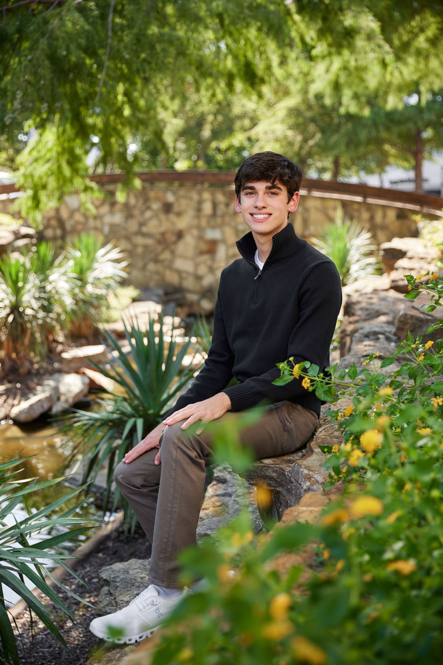 Smiling teenage boy sits on rocks near a garden pond surrounded by lush greenery and flowers.