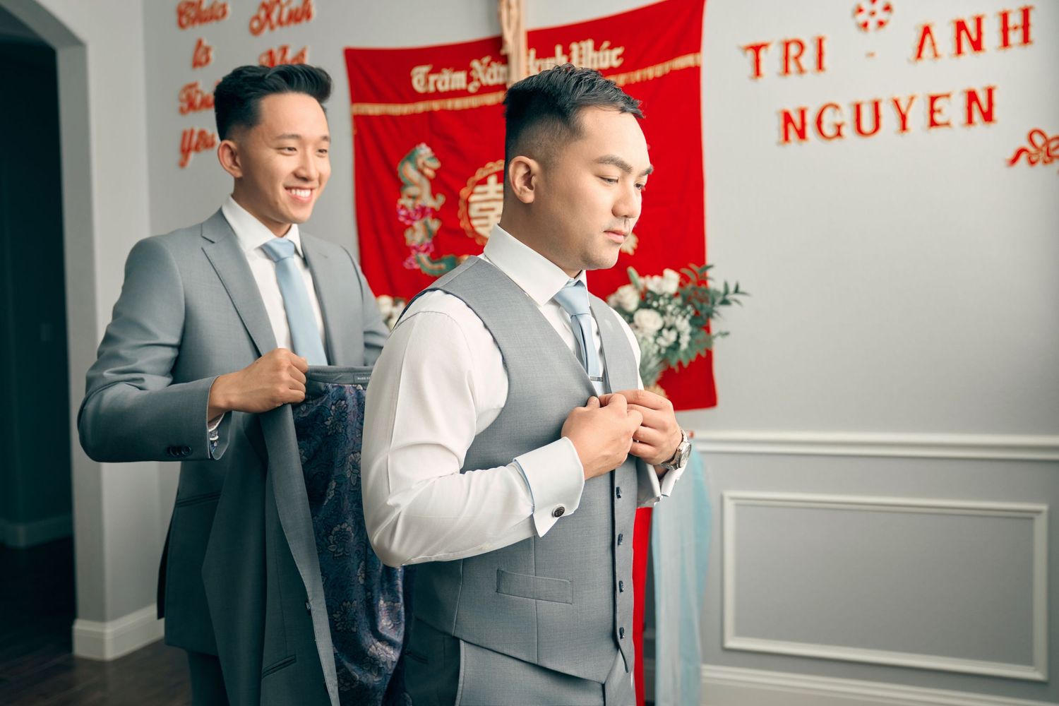 Groom getting dressed in grey suit with groomsman assistance before Vietnamese wedding ceremony.